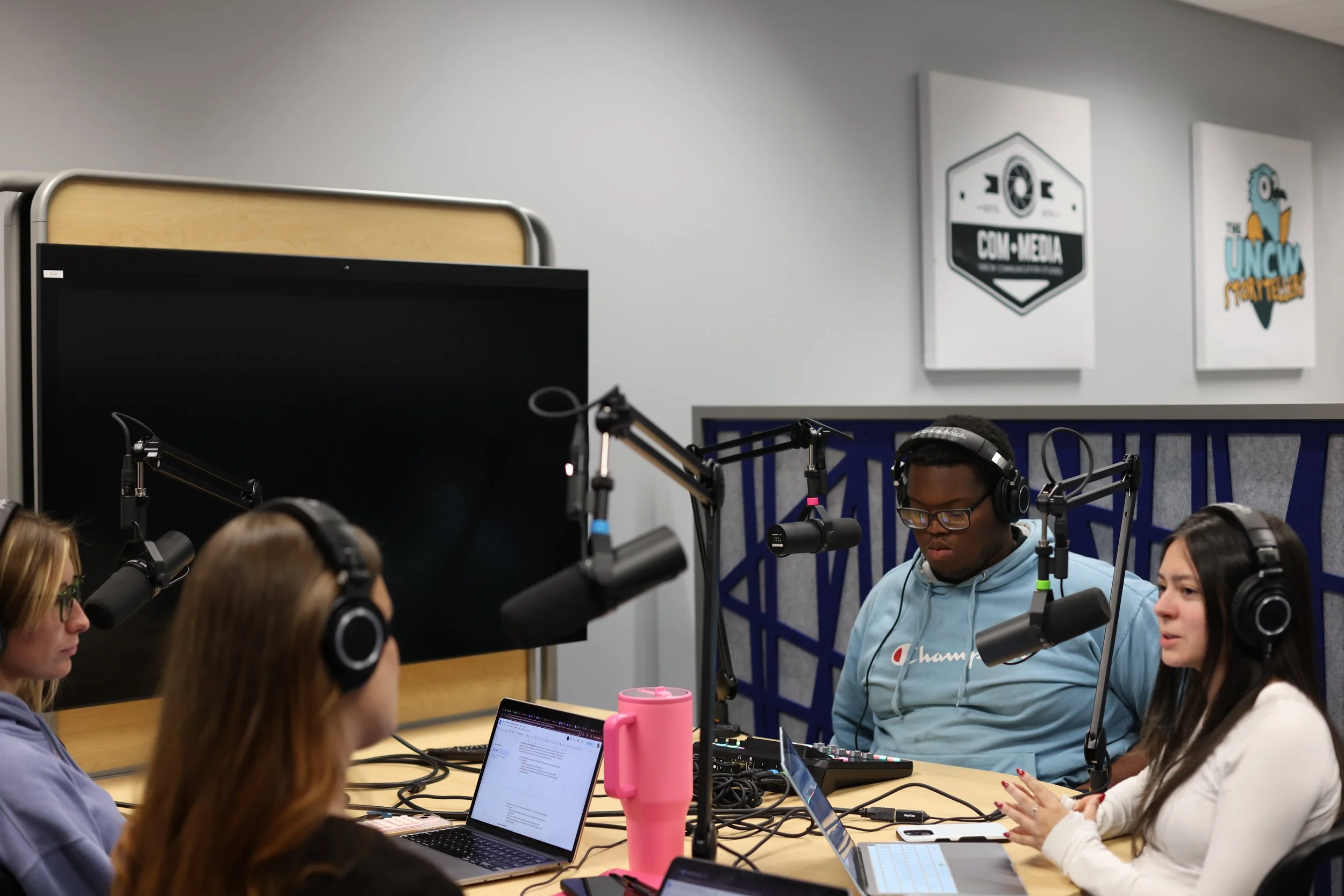 Four people sitting around a table with microphones, laptops, and headphones in a recording studio or podcast setting.