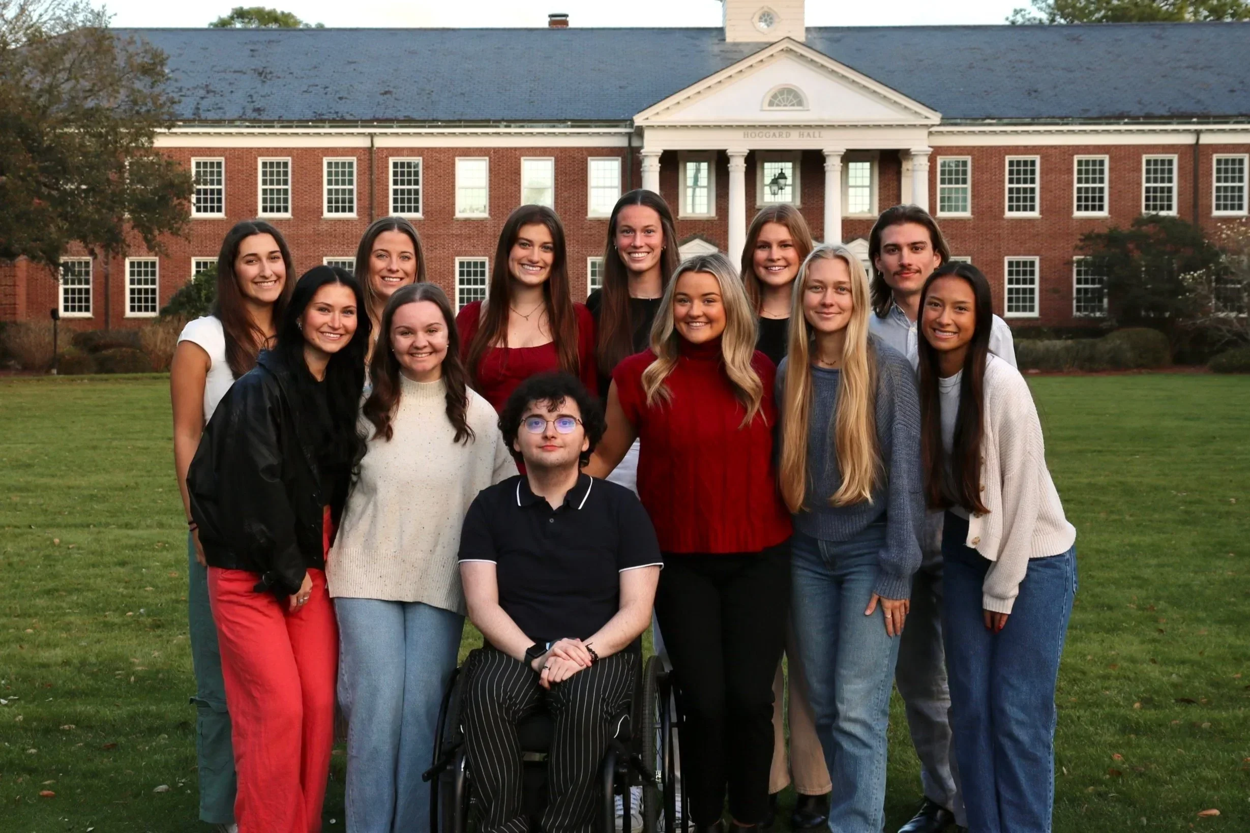 Group of young people, including a person in a wheelchair, standing on a grassy lawn in front of a brick building with columns. The group is smiling and diverse in appearance.