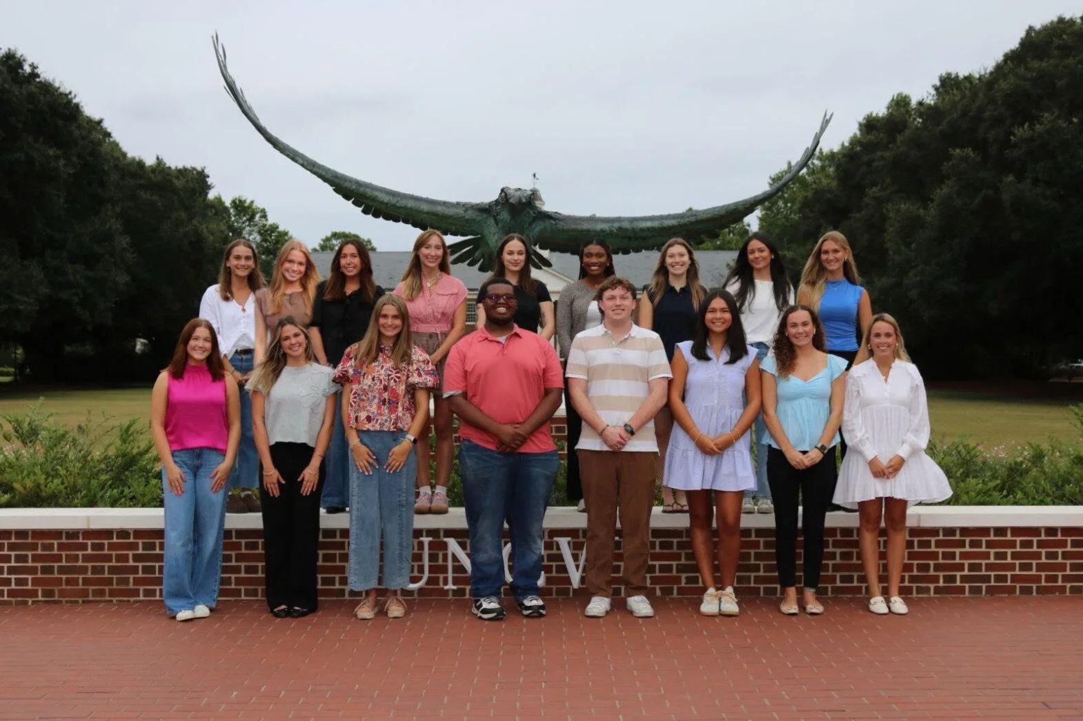 Group of 16 young adults posing in front of a large eagle sculpture on a college campus.