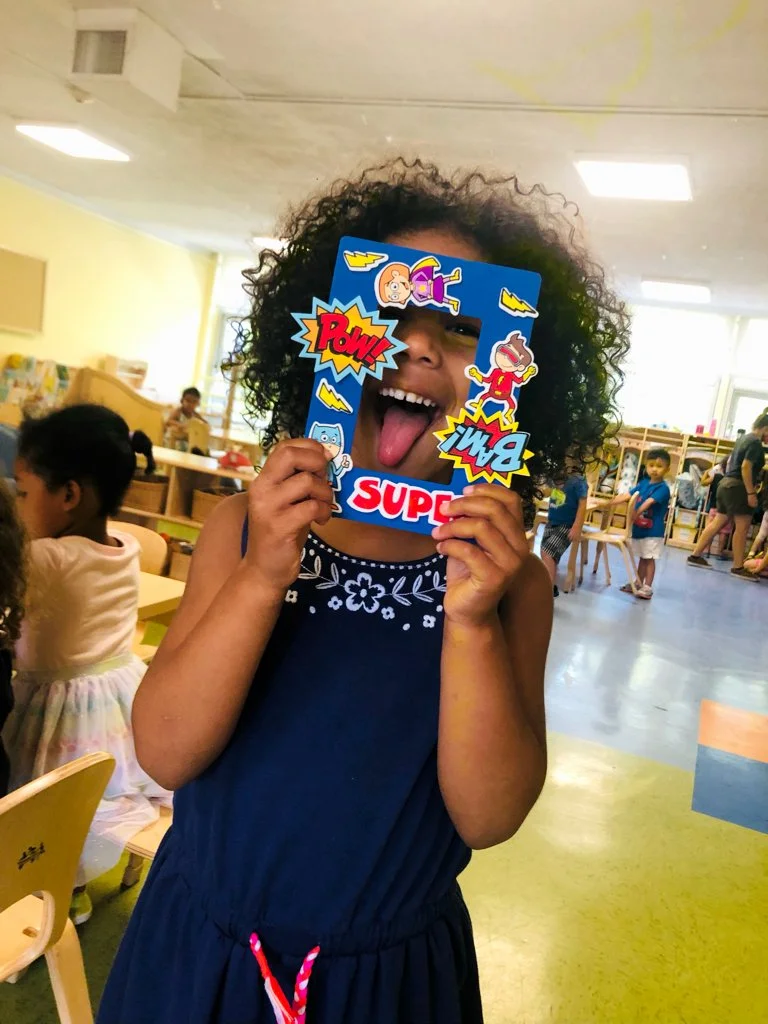 A young girl with curly hair holding a superhero-themed photo frame covering her eyes and part of her face, sticking out her tongue in a classroom or daycare setting.