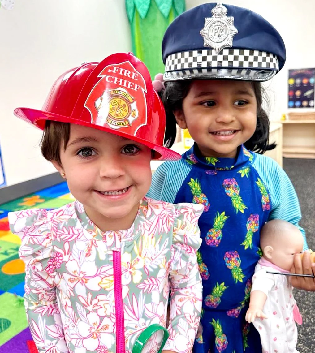 Two young girls dressed as a fire chief and police officer, wearing firefighter and police hats, smiling, with one girl holding a doll.