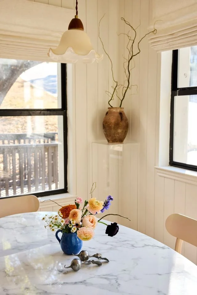 A white marble table with a blue vase filled with colorful flowers, two small decorative silver objects, and a black flower. The room has white paneling, two windows with blinds, and a pendant light hanging above. There is a large brown vase on a wall shelf with tall twisted branches inside.