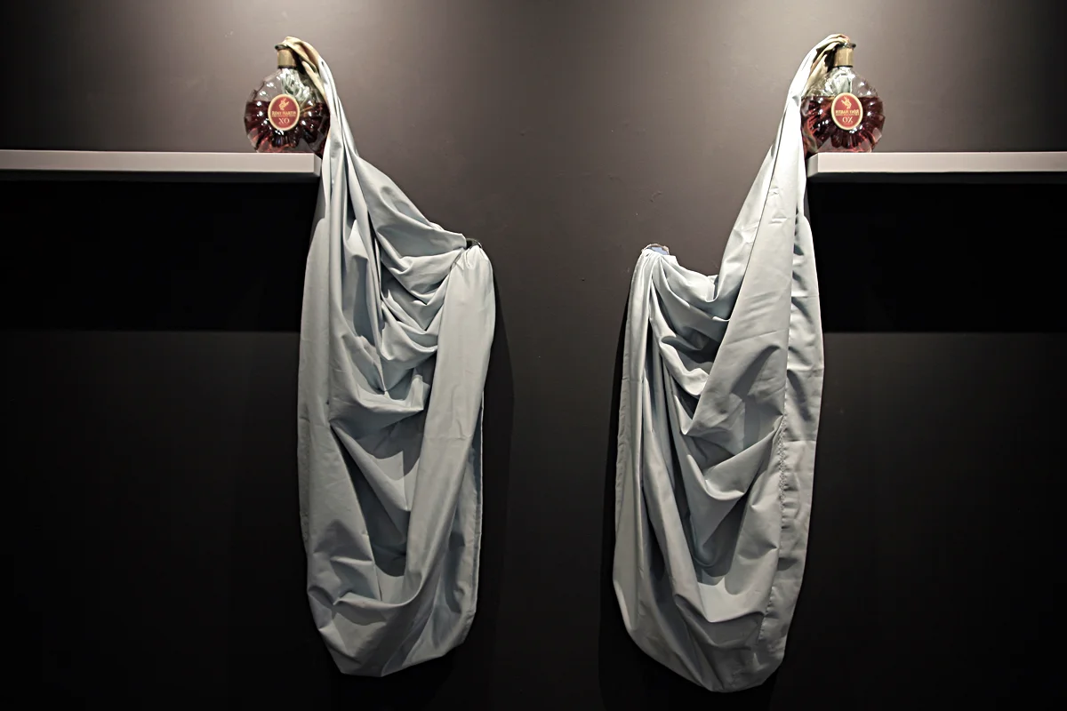 SWEAT (1) , 2009, Two Remy Martin XO, bed sheet and two shelves, Dimension variable. In this work one bed sheet connects two bottles of Remy Martin XO Cognac, traveling from one bottle to the next through two holes in the gallery wall, the bed sheet travels behind the gallery wall.