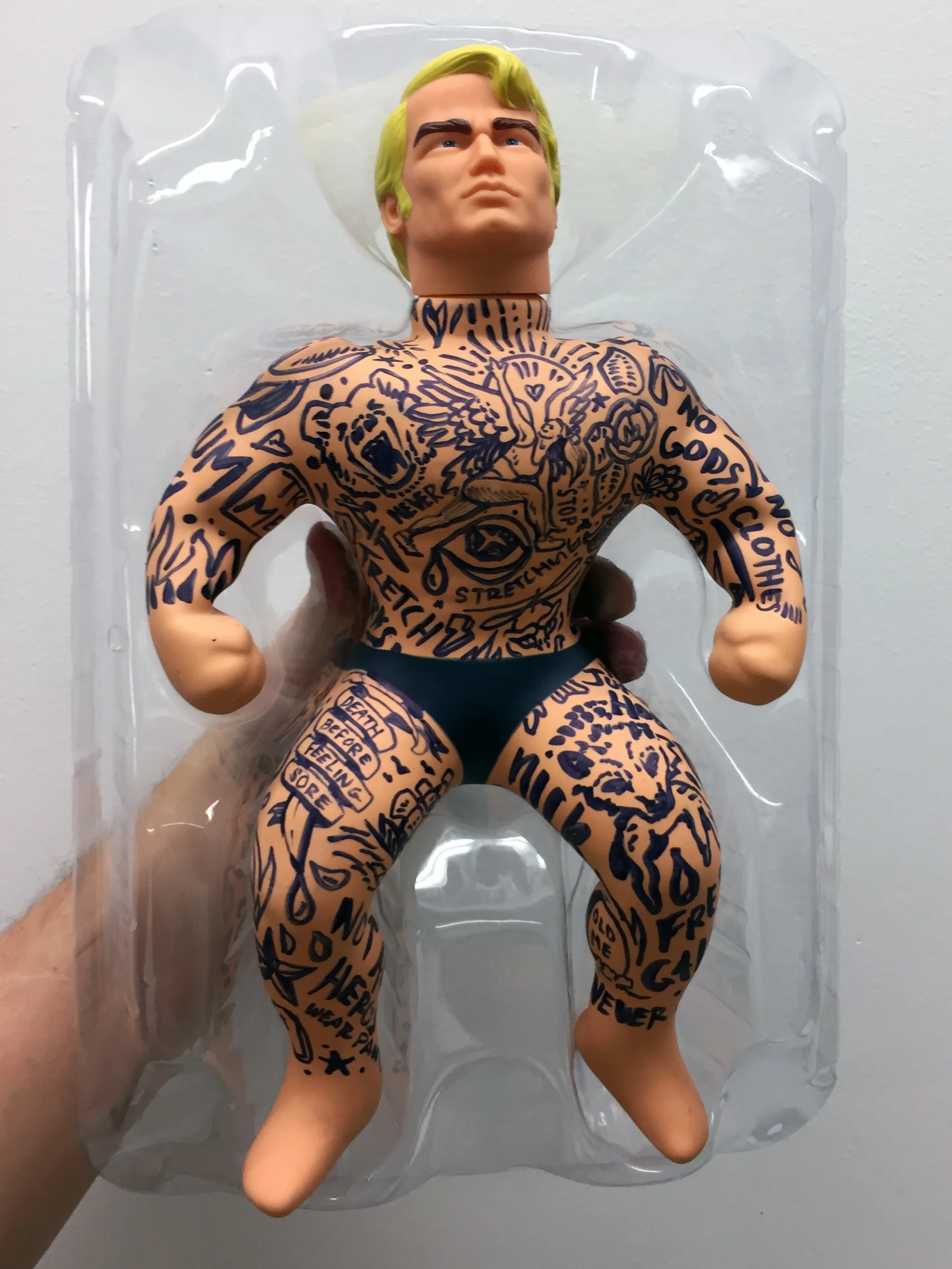 A toy action figure with a muscular build and a serious expression, covered in black tattoo-style drawings and texts on its arms, chest, and legs, packaged in clear plastic.