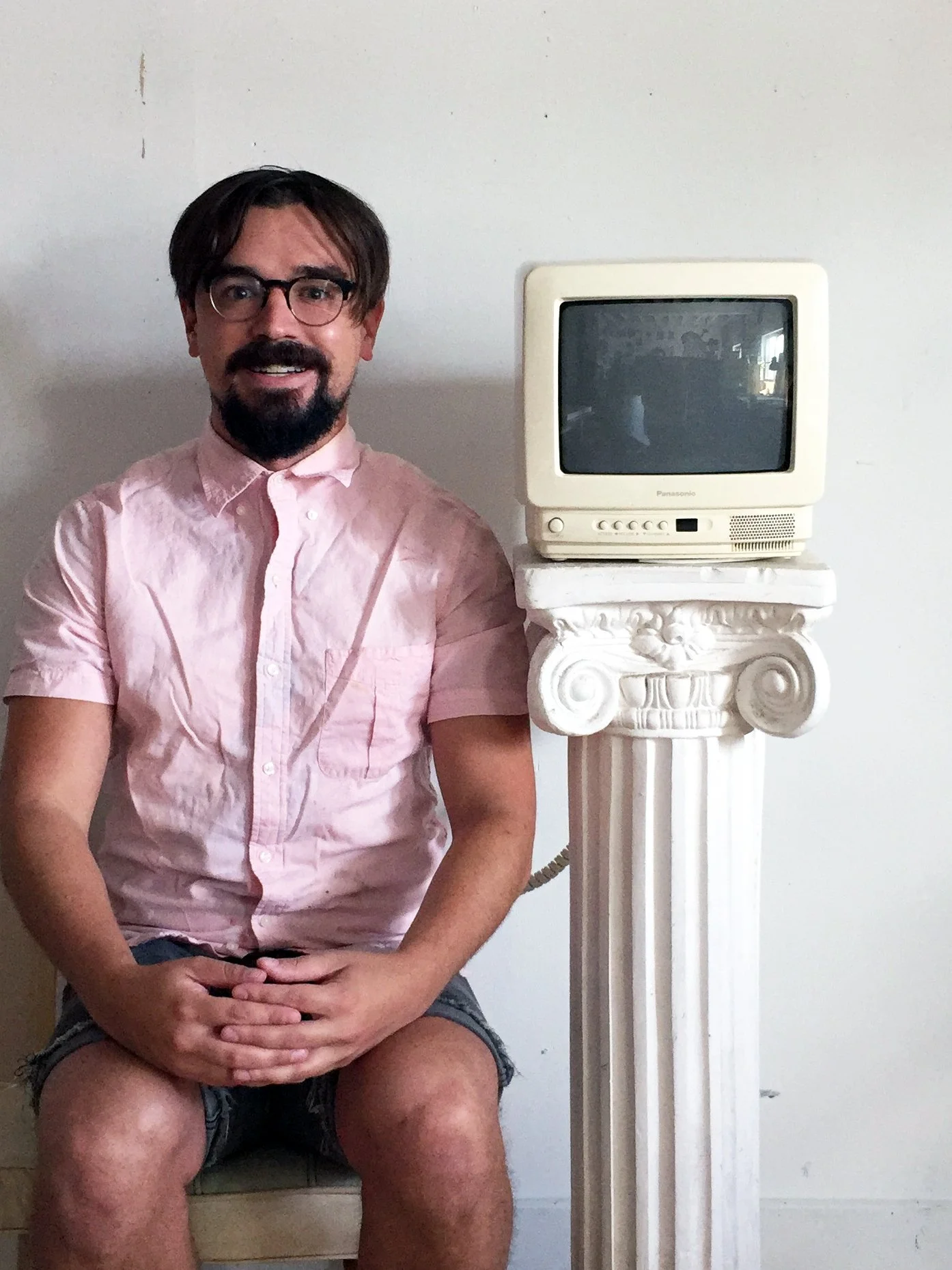 A man with glasses, a beard, and brown hair is sitting on a chair, smiling, wearing a pink shirt and shorts. Next to him is an old, small CRT television on a white, decorative column.