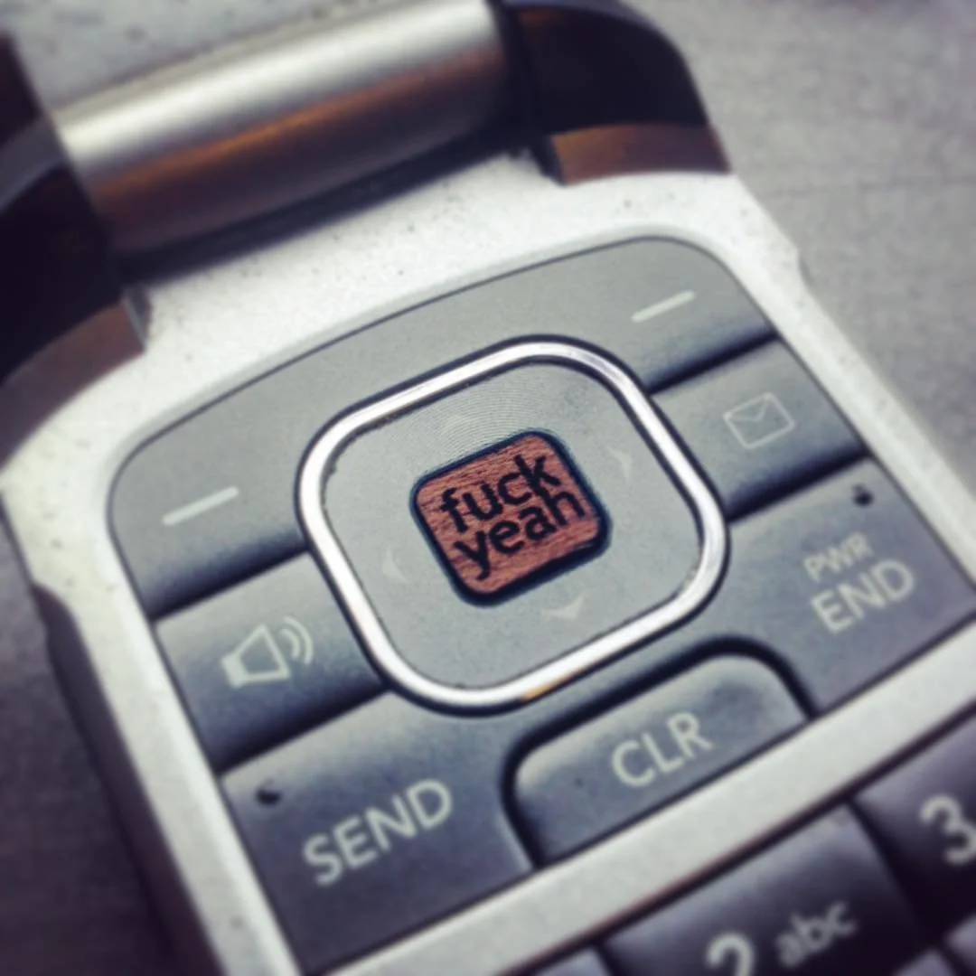 A cellphone with a small digital screen displaying the words 'FUCK YEAH' in black text on an orange background.