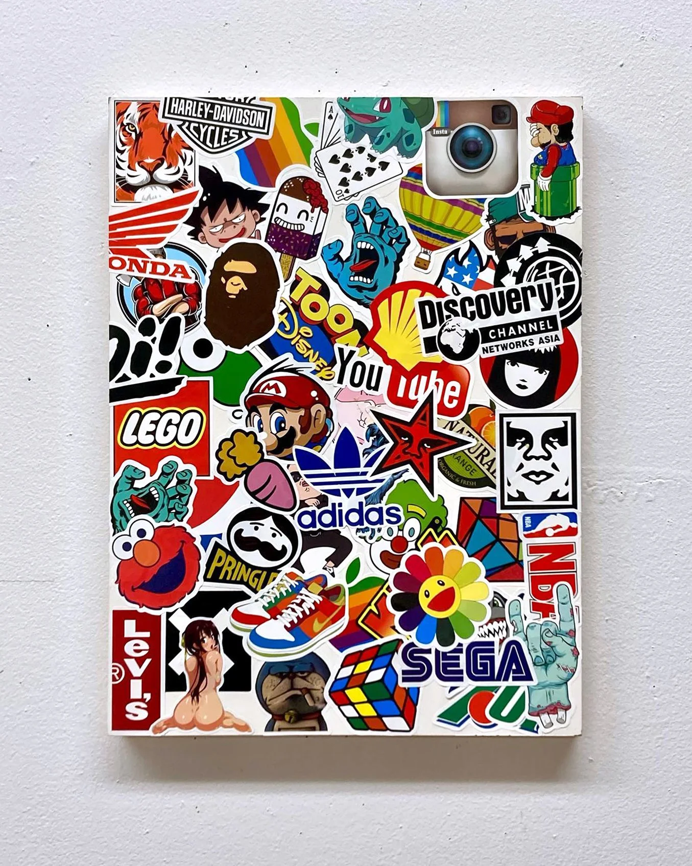 A white wall with a large rectangular collage of various colorful stickers, including brand logos like Harley Davidson, Lego, Adidas, SEGA, Levi's, YouTube, Discovery Channel, and Nintendo, as well as cartoon characters, a Rubik's Cube, and other graphical stickers.
