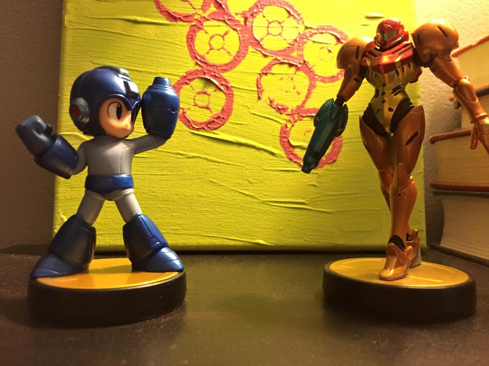 Mega Man (left) and Samus (right) amiibo figures face off in front some of homemade artwork, courtesy of my wife. They might both have arm cannons and be from the future, but they're quite different heroes.