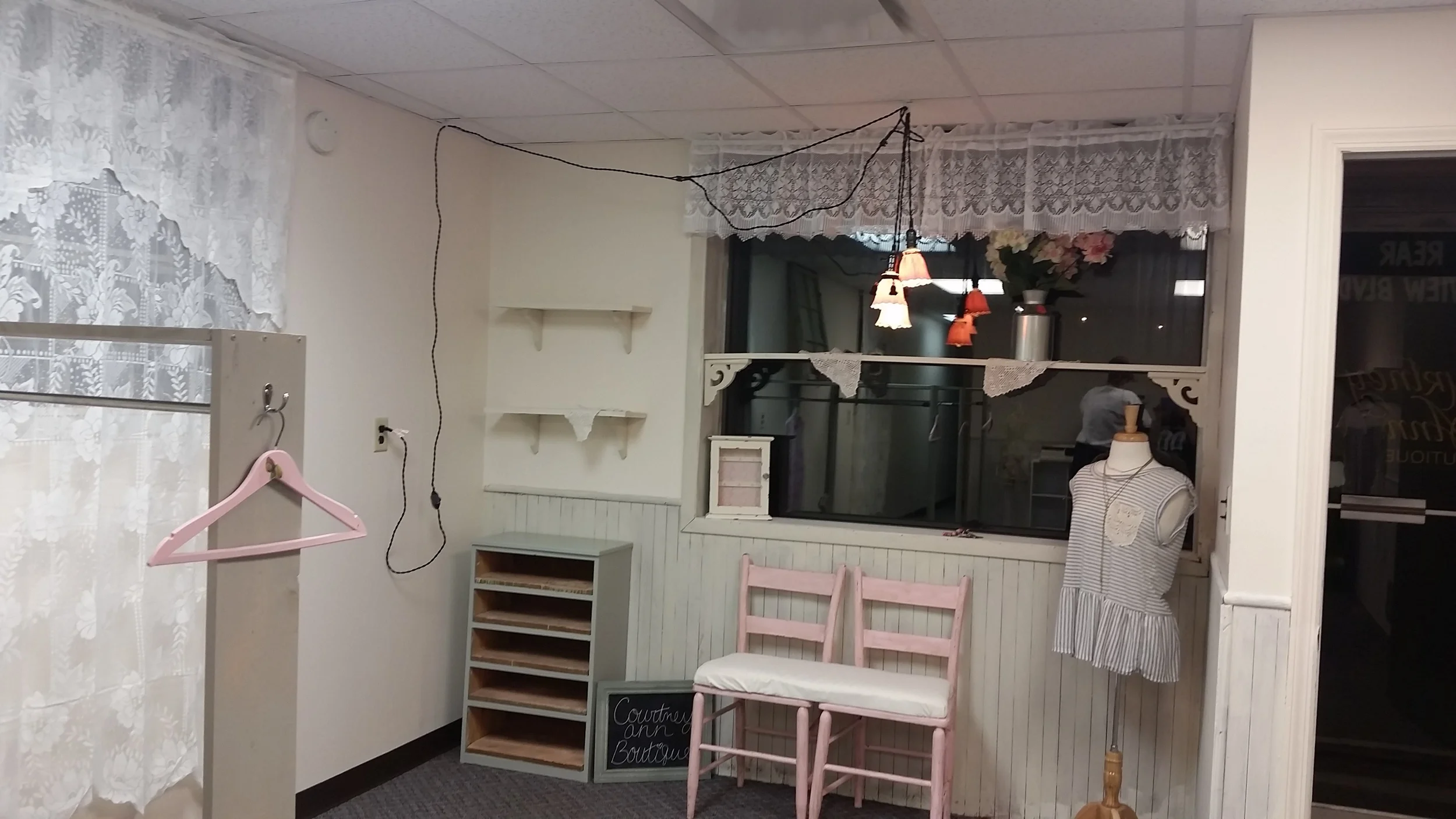 Kristen wanted a coffee bar, so a repurposed dresser and some shelves filled the bill. Reimagined chairs painted a pretty pink provide a place for waiting and give the vintage boutique vibe the owners wanted. Lace helped the windows make the transition. Pinks and grays make the carpet make sense in this space. The paneling got whitewashed to provide that shabby feel.