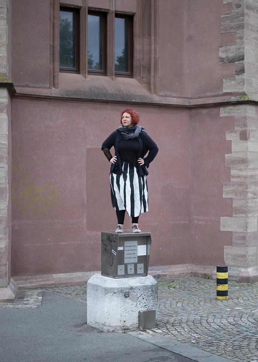 Ladeninhaberin Gabriela Strasser Steht auf einer Säuel. Sie trägt ein schwarz-weiss gestreiften Rock, schwarzen Oberteil und hat rote Haare und roten Lippenstift. Der Hintergrund ist eine Sandstein farbige Wand.