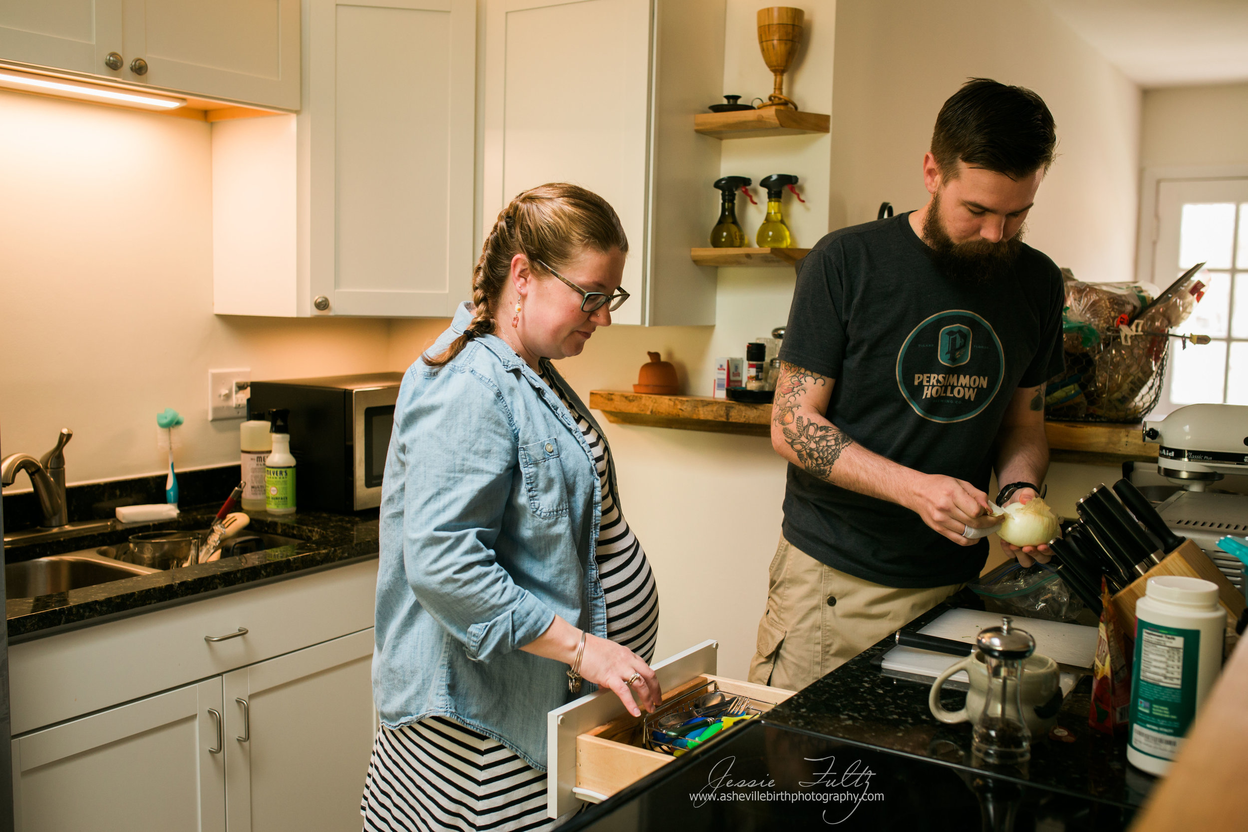 redheaded pregnant woman in striped dress looking in utensil drawer while husband peels an onion