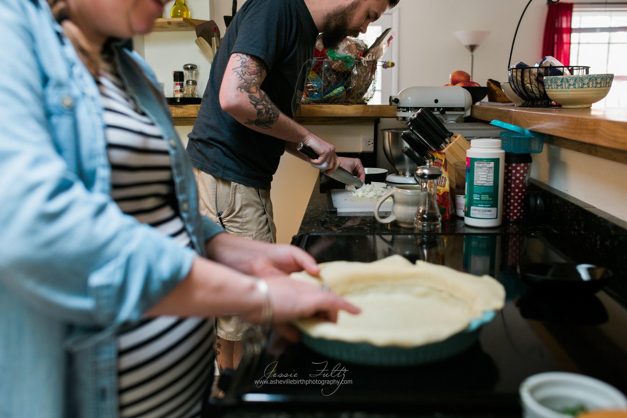 man in black shirt chopping onions while pregnant wife makes pie crust in the foreground