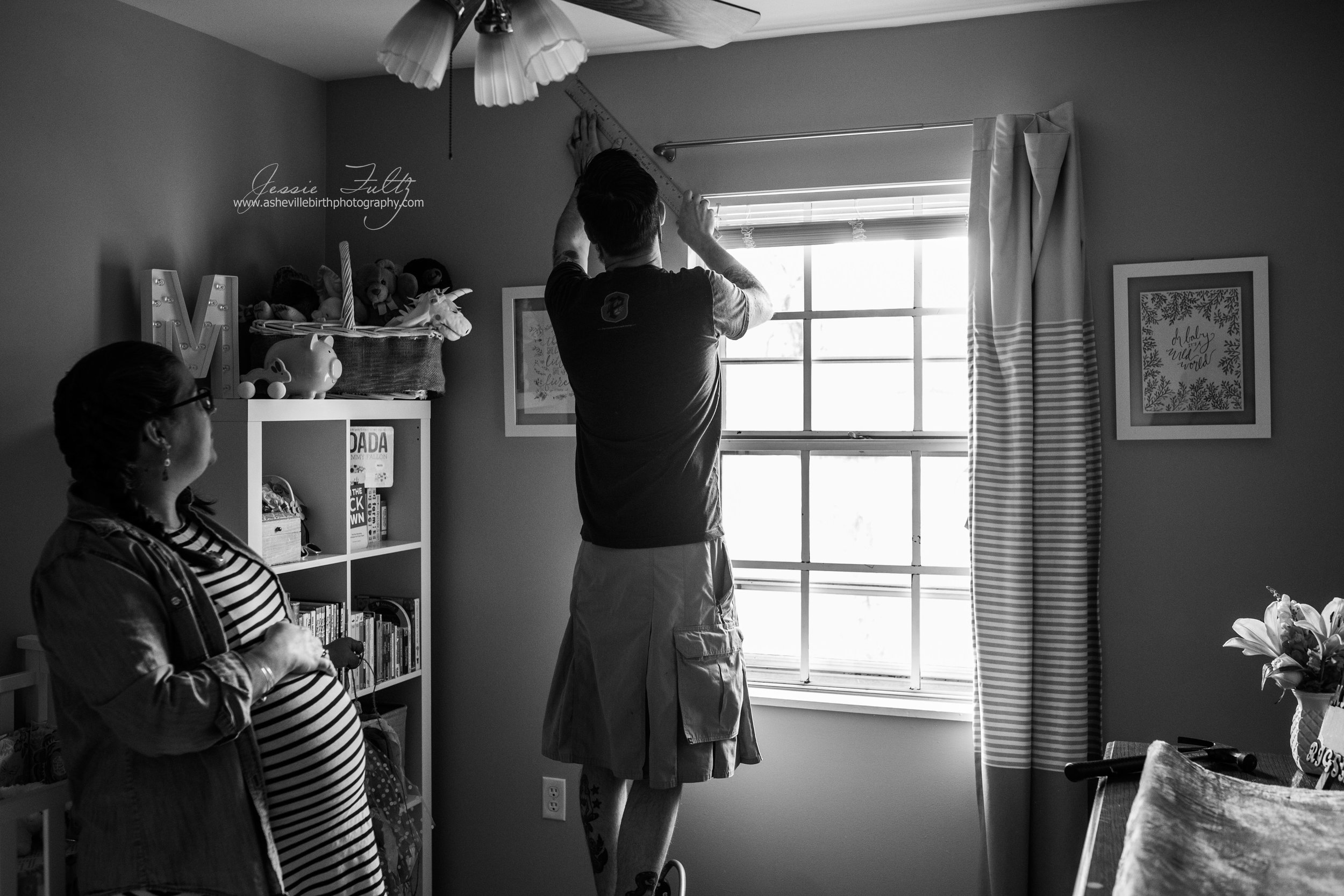black and white picture of a pregnant woman holding her belly and watching her husband who is wearing a kilt measure the wall in their nursery