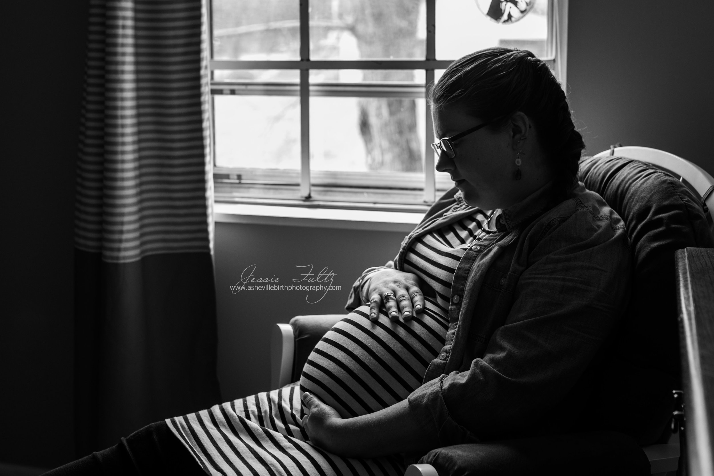 black and white picture of pregnant woman looking down at her belly sitting in a glider chair next to the window in her nursery