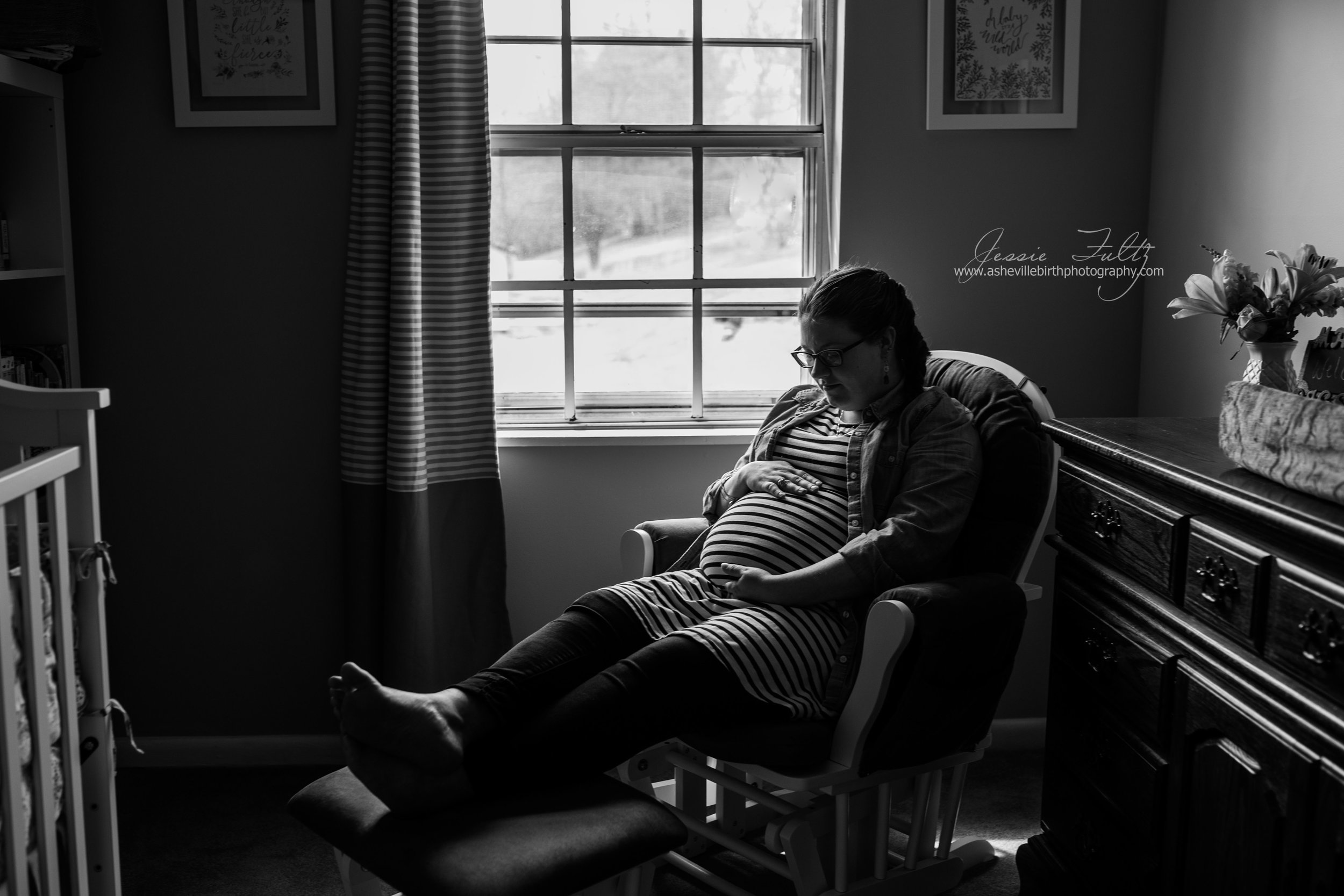 wide-angle black and white picture of pregnant woman in striped dress looking down at her belly sitting in a glider chair next to the window in her nursery