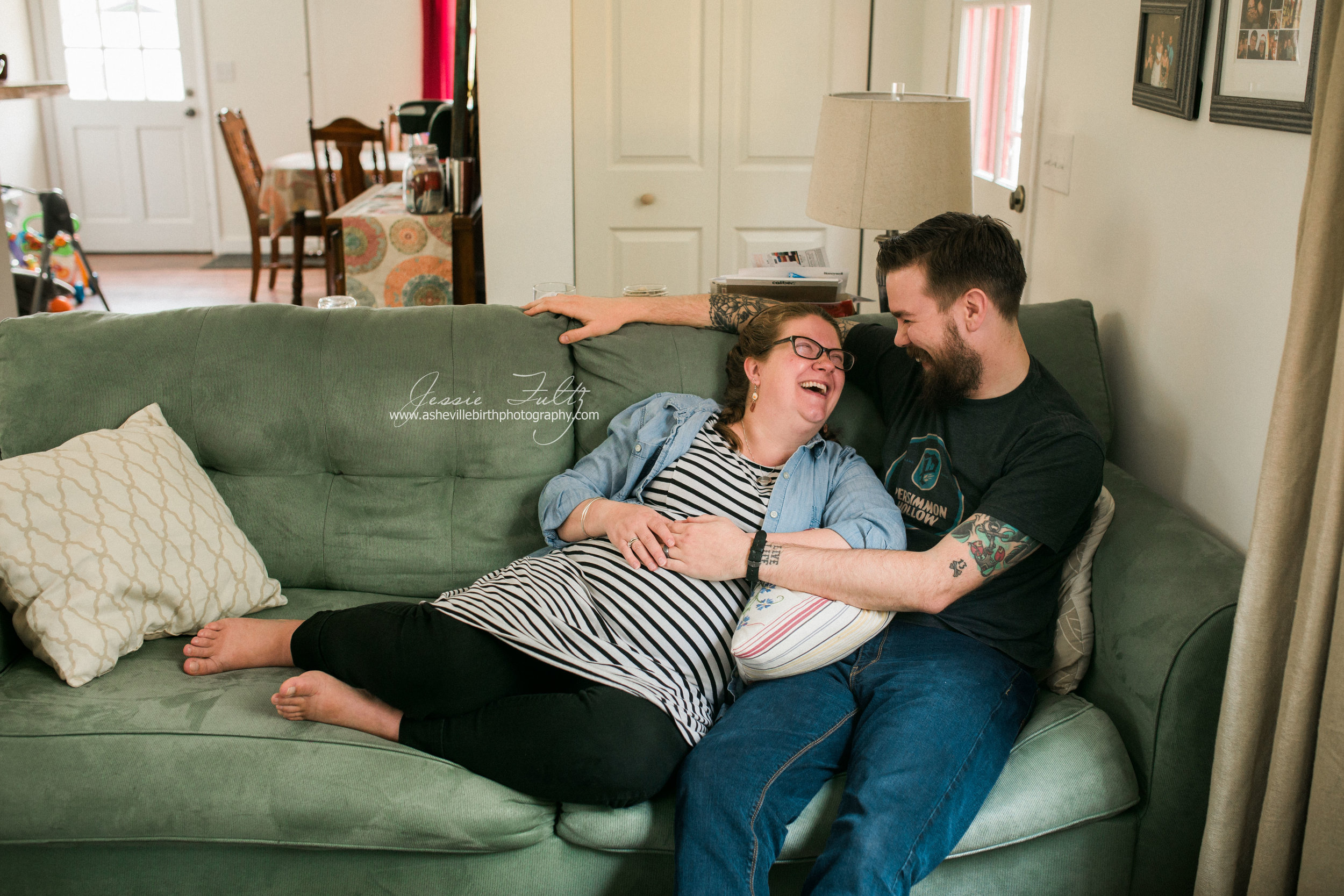 pregnant woman in striped dress and husband cuddling up on the couch and laughing