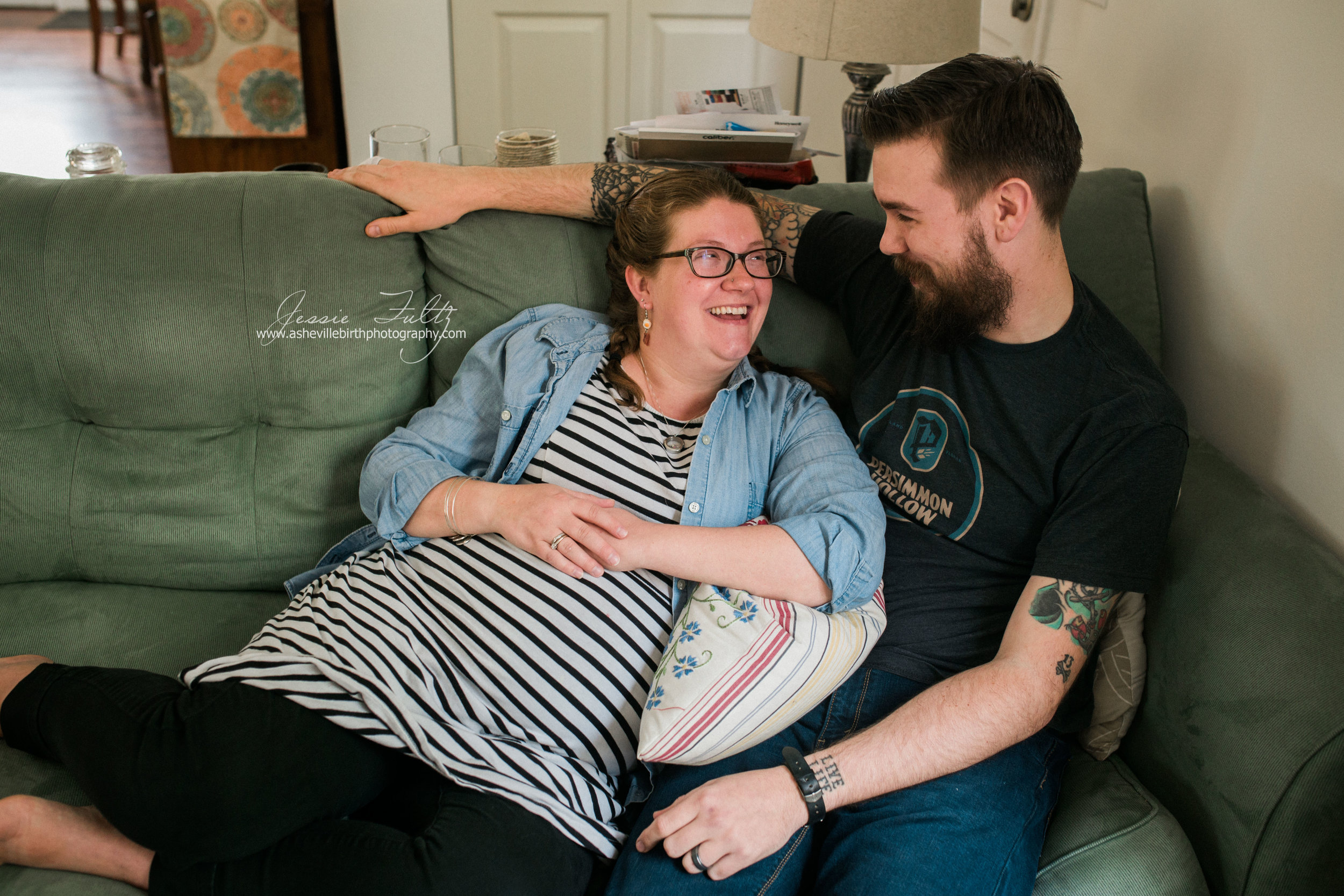 pregnant woman in striped dress and husband cuddling up on the couch and laughing