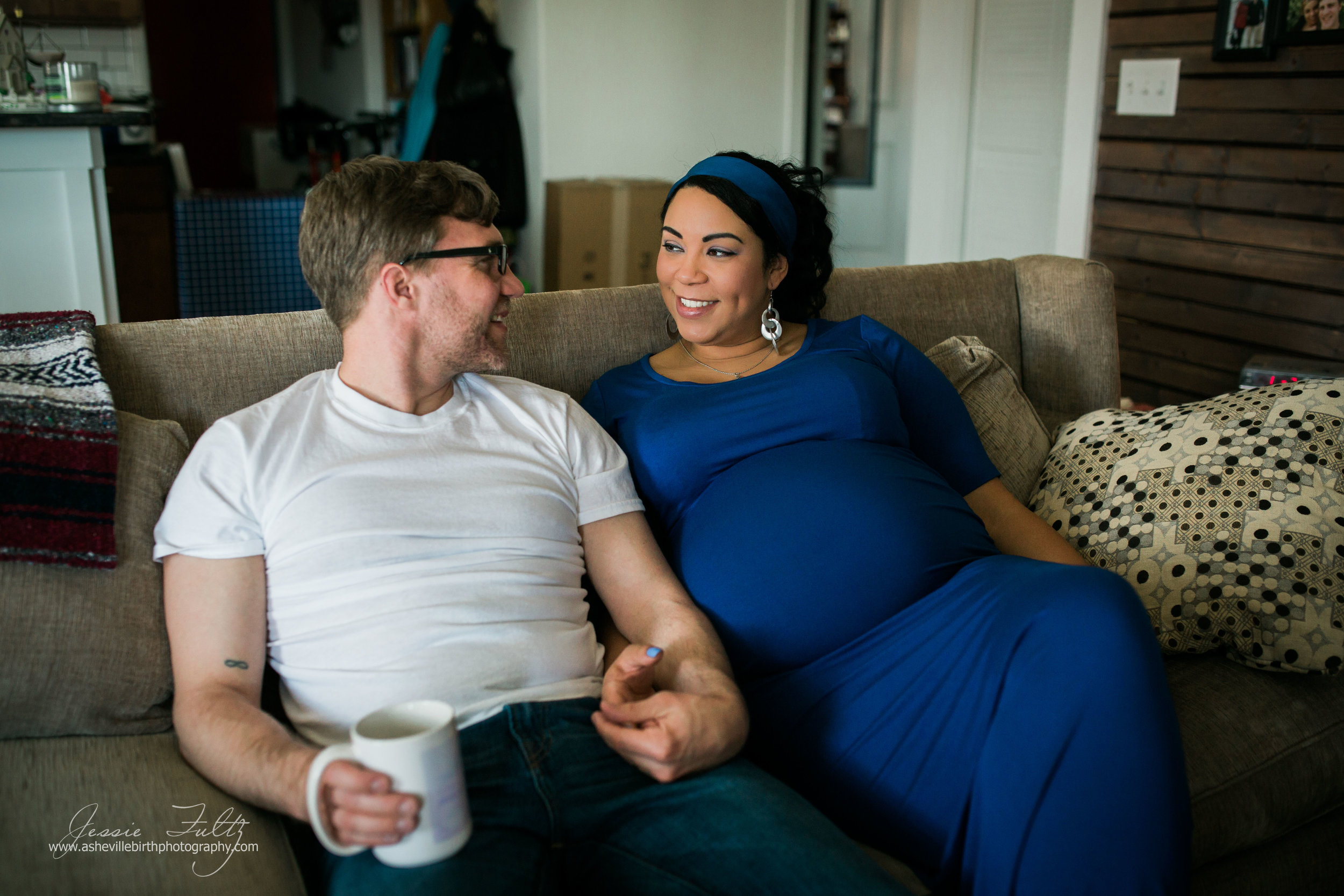 pregnant woman in blue dress and husband sitting on couch holding hands and smiling at each other