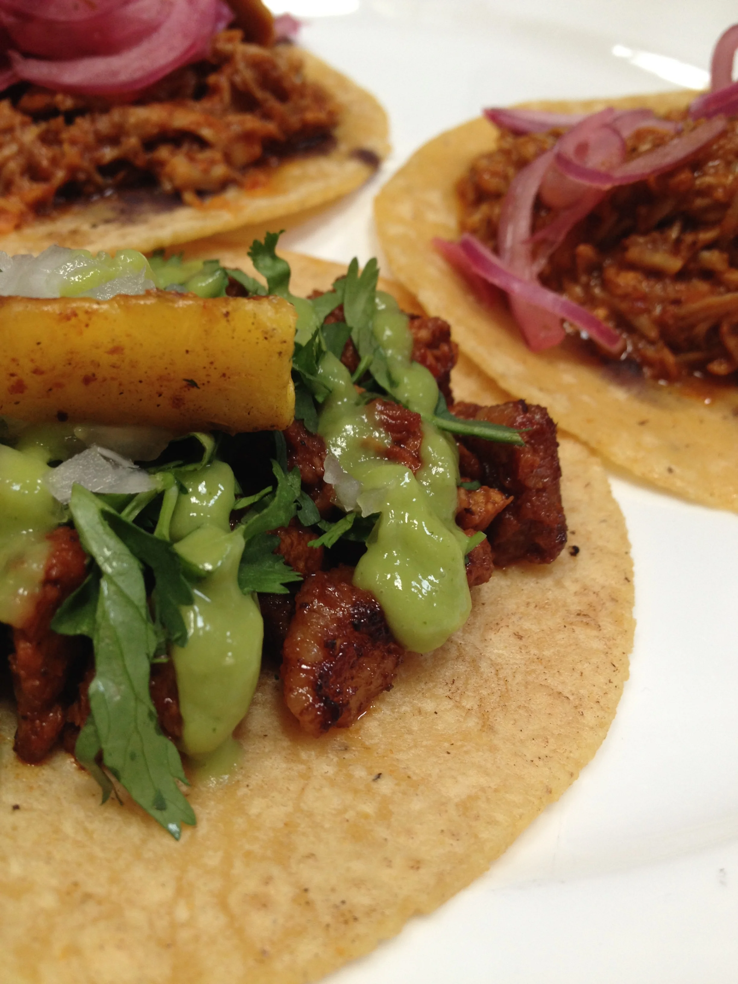 (foreground) Al Pastor taco with adobo-chile rubbed pork shoulder, pineapple, and avocado (background) Two Cochinita Pibil tacos with pulled pork shoulder, pickled red onion, oregano, habanero, and black beans