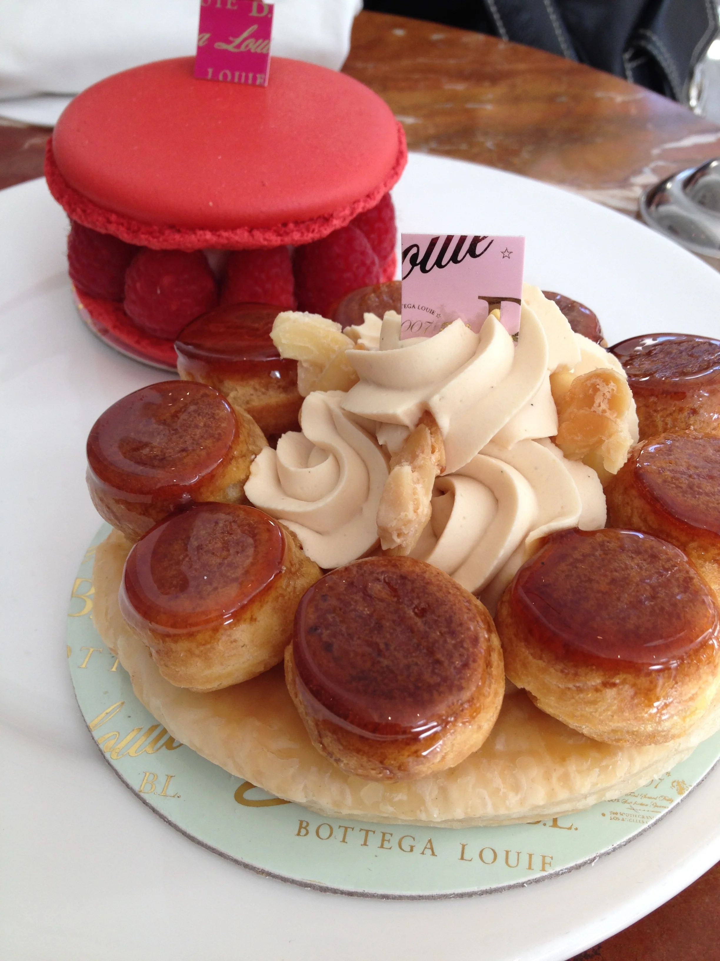 (Background) Le Grand Macaron, a French almond cookie with vanilla cream, raspberry jam, and raspberries (Foreground) St. Honoré cake with a base of puff pastry, cream puffs, and a ring of piped pâte à choux.