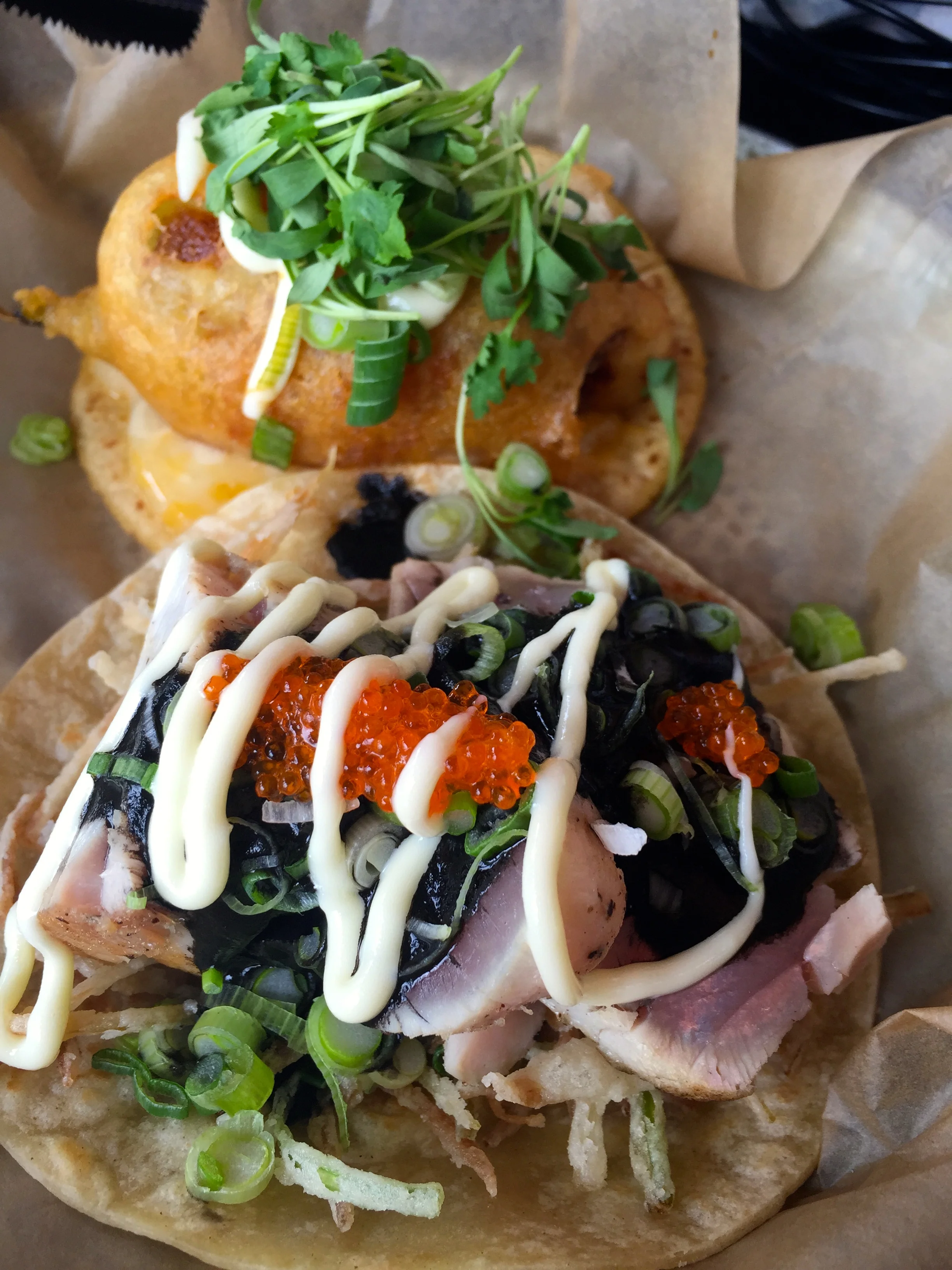 (Foreground) Taco of the week, with Albacore Tuna (medium rare), on a bed of fried leeks, topped with squid ink, habanero sauce, wasabi aioli, scallion & tobico roe on a flour tortilla.(Background) Sea Urchin Chile Guero Taco with tempura batter…