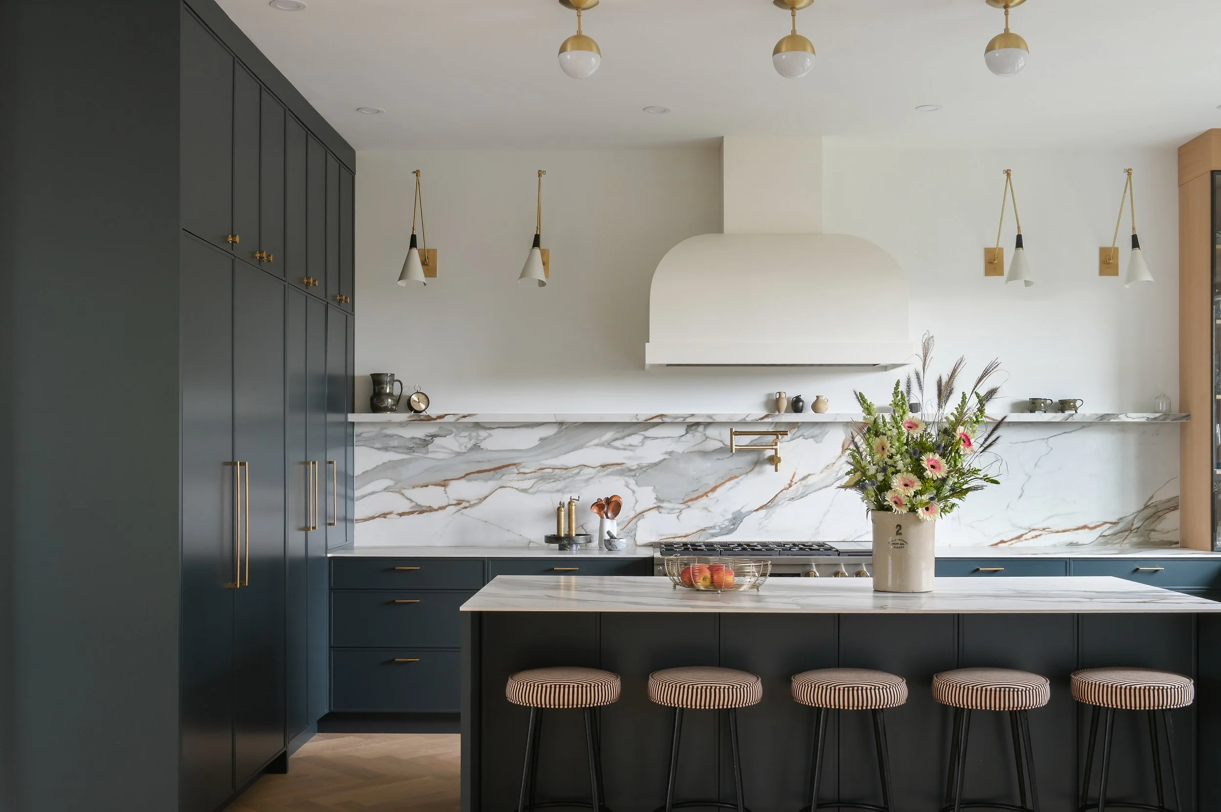 Luxury custom kitchen by Accentrix Design featuring dark cabinetry, dramatic marble backsplash, oversized waterfall-style island, brass accents, and statement range hood.