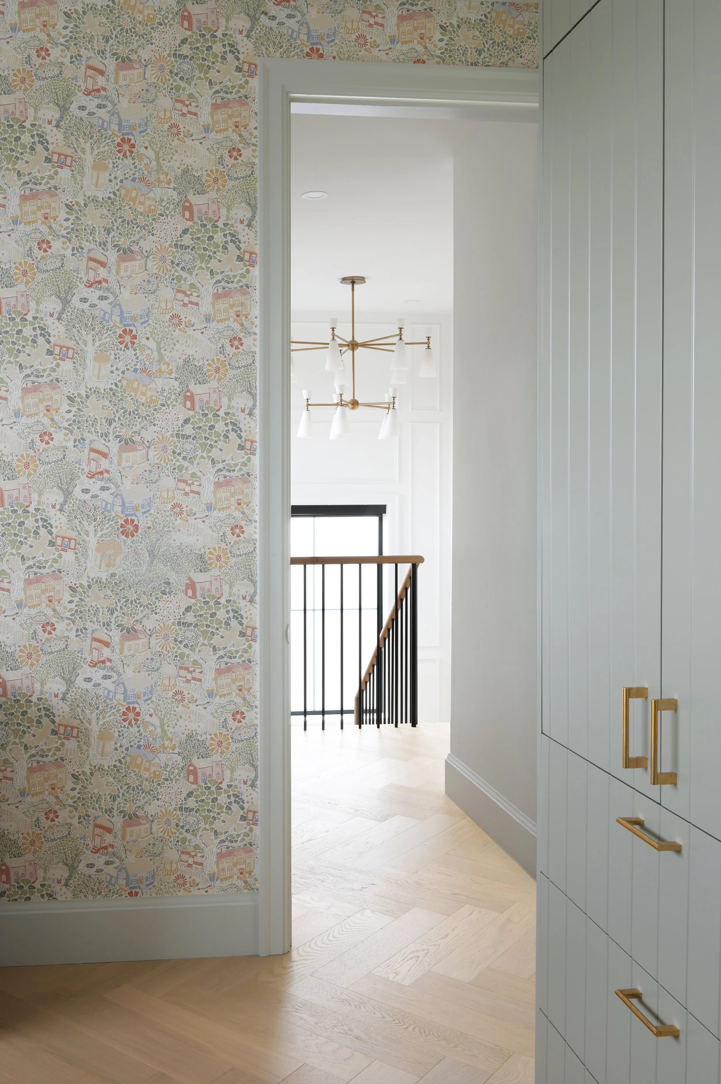 Custom residential interior by Accentrix Design featuring whimsical patterned wallpaper, soft sage green built-in cabinetry, brushed brass hardware, and light herringbone hardwood flooring.