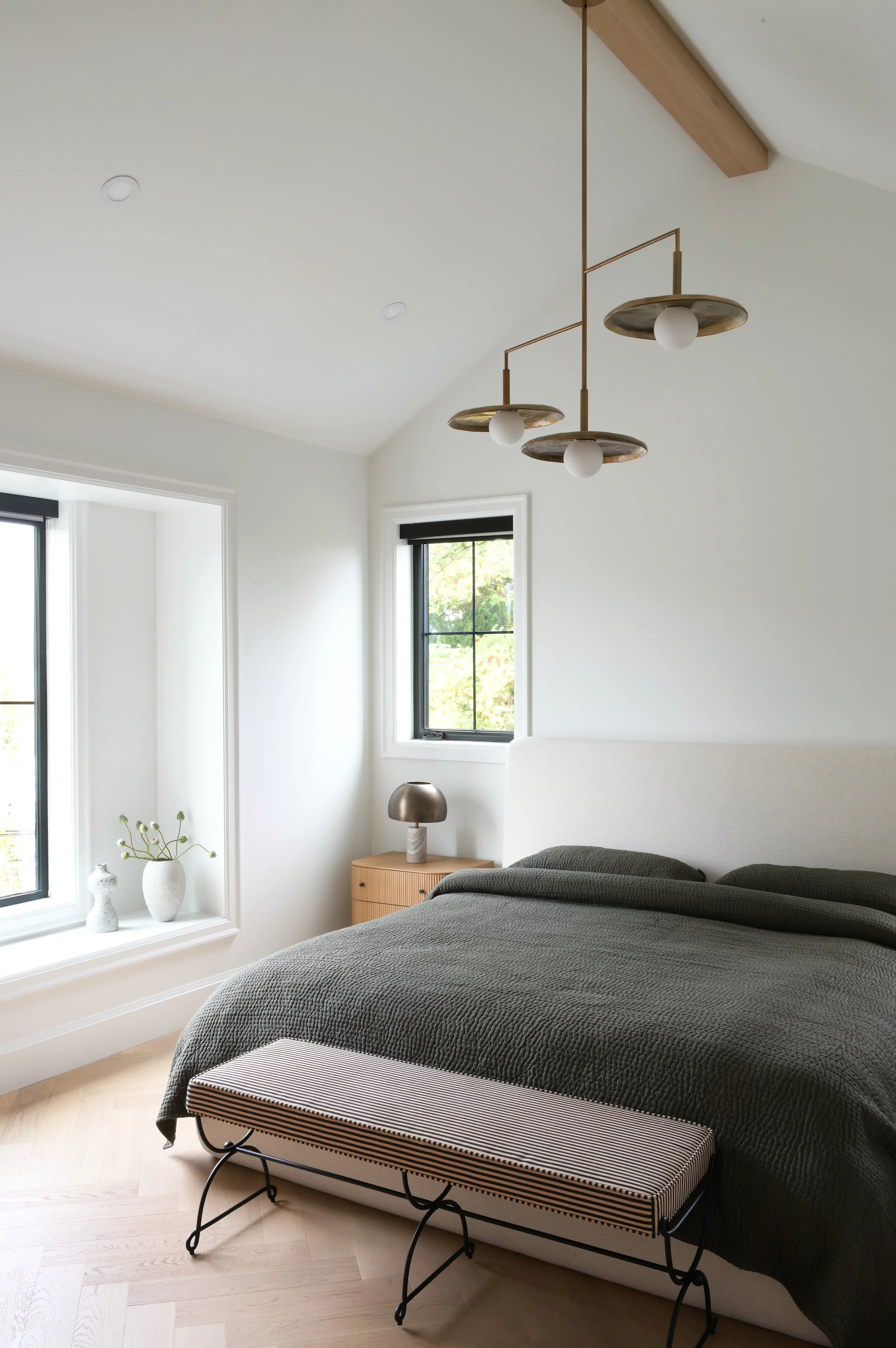 Modern luxury bedroom by Accentrix Design featuring a curved upholstered headboard, charcoal bedding, brass statement pendant lighting, natural wood nightstand, and light herringbone hardwood floors.