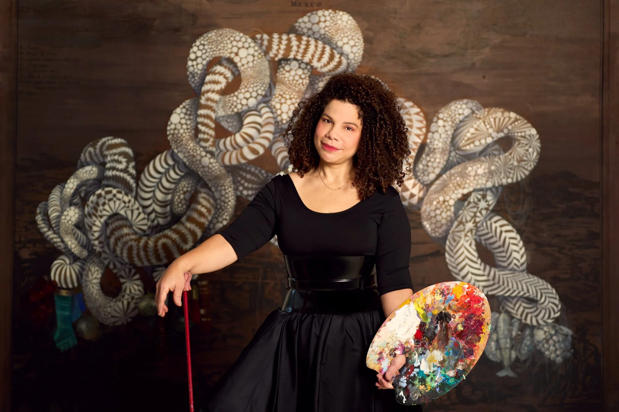 Editorial portrait of painter Firelei Báez in her studio, holding a palette before her large patterned painting, photographed by Christopher Garcia Valle