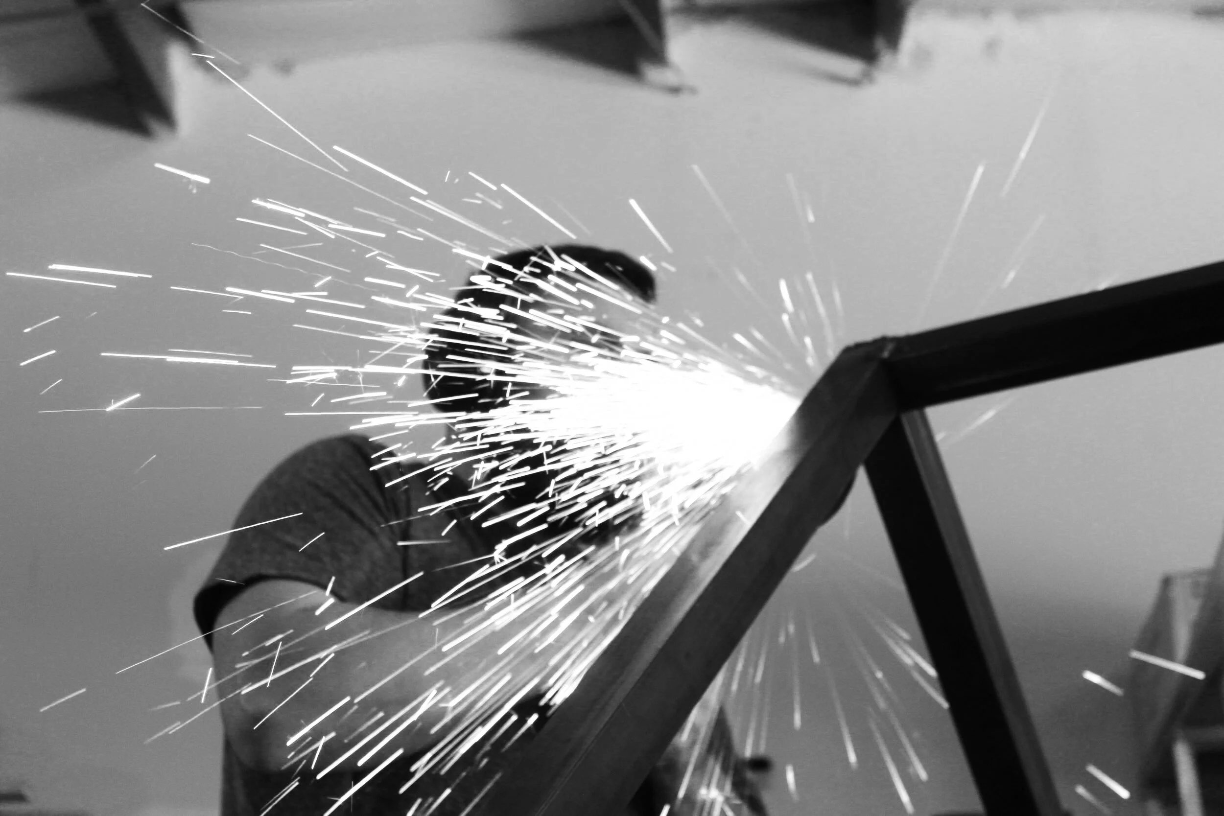 Black and white photo of sparks flying from metal grinding or welding work on a piece of metal, with a hand wearing a glove holding the metal piece.