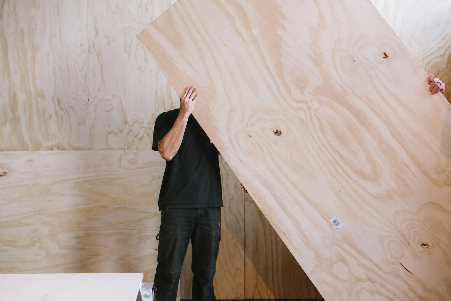 Person holding a large piece of plywood in a room with plywood walls.