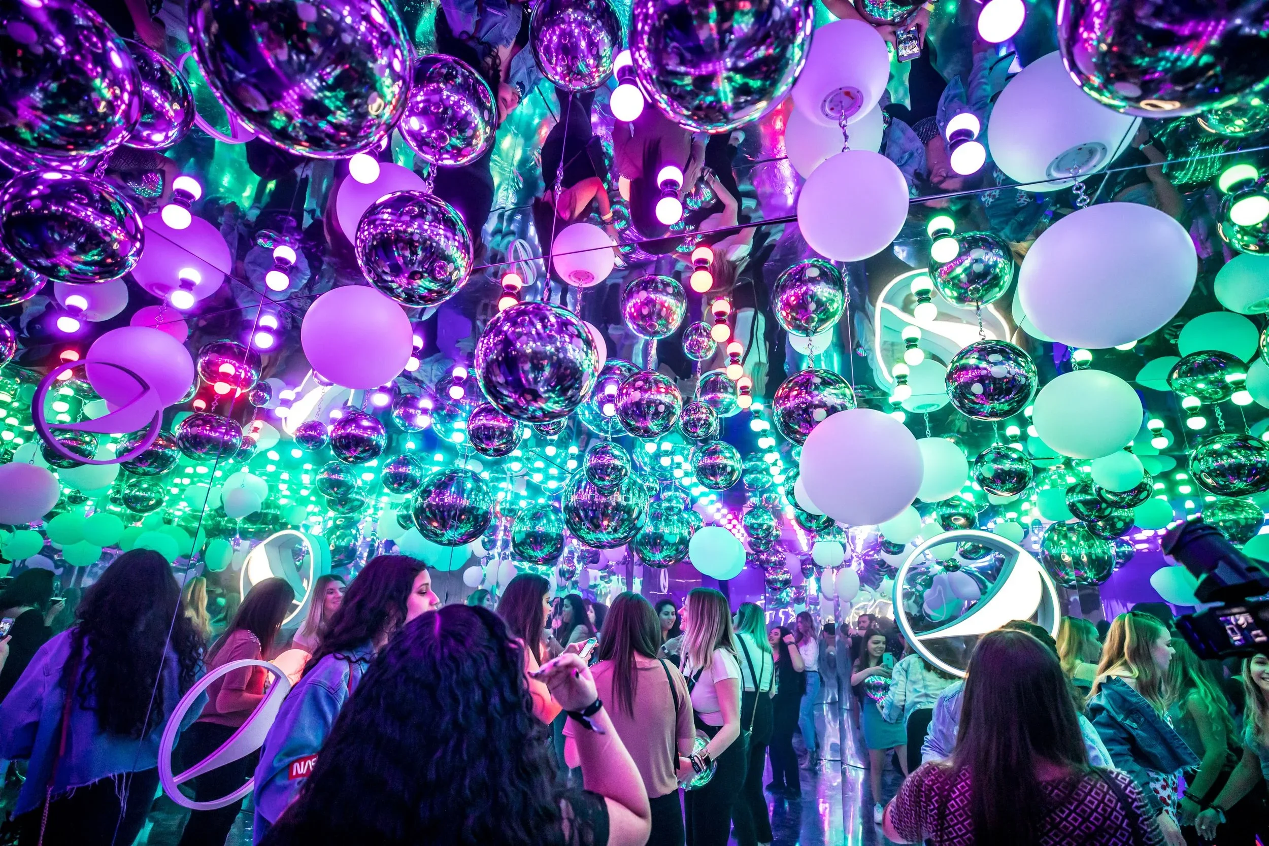 Immersive event installation with mirrored ceiling, hanging spheres, branded elements, and colorful lighting.