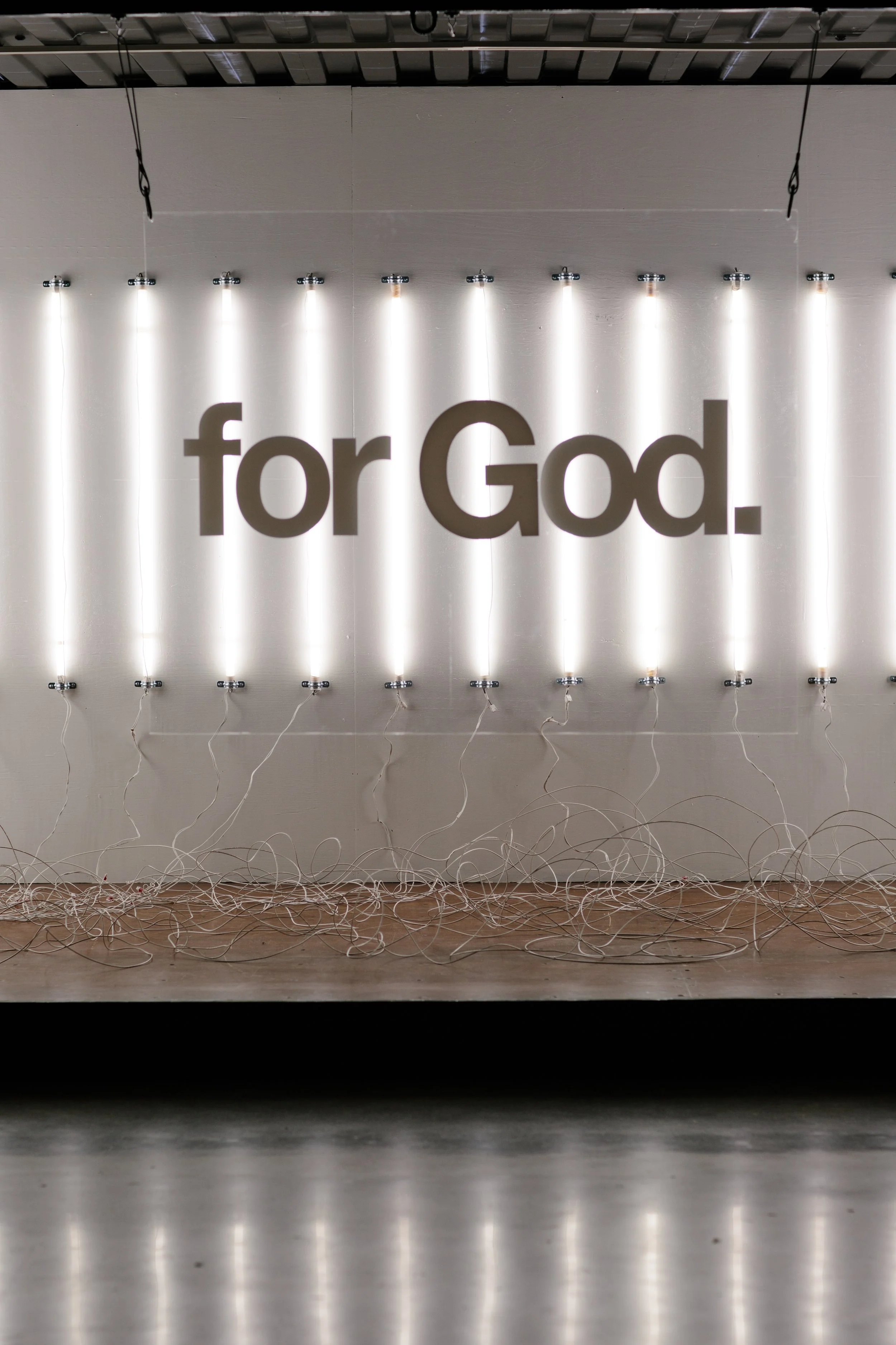 White wall with illuminated vertical light tubes and large text 'for God.' in the center, with tangled wires on the floor.