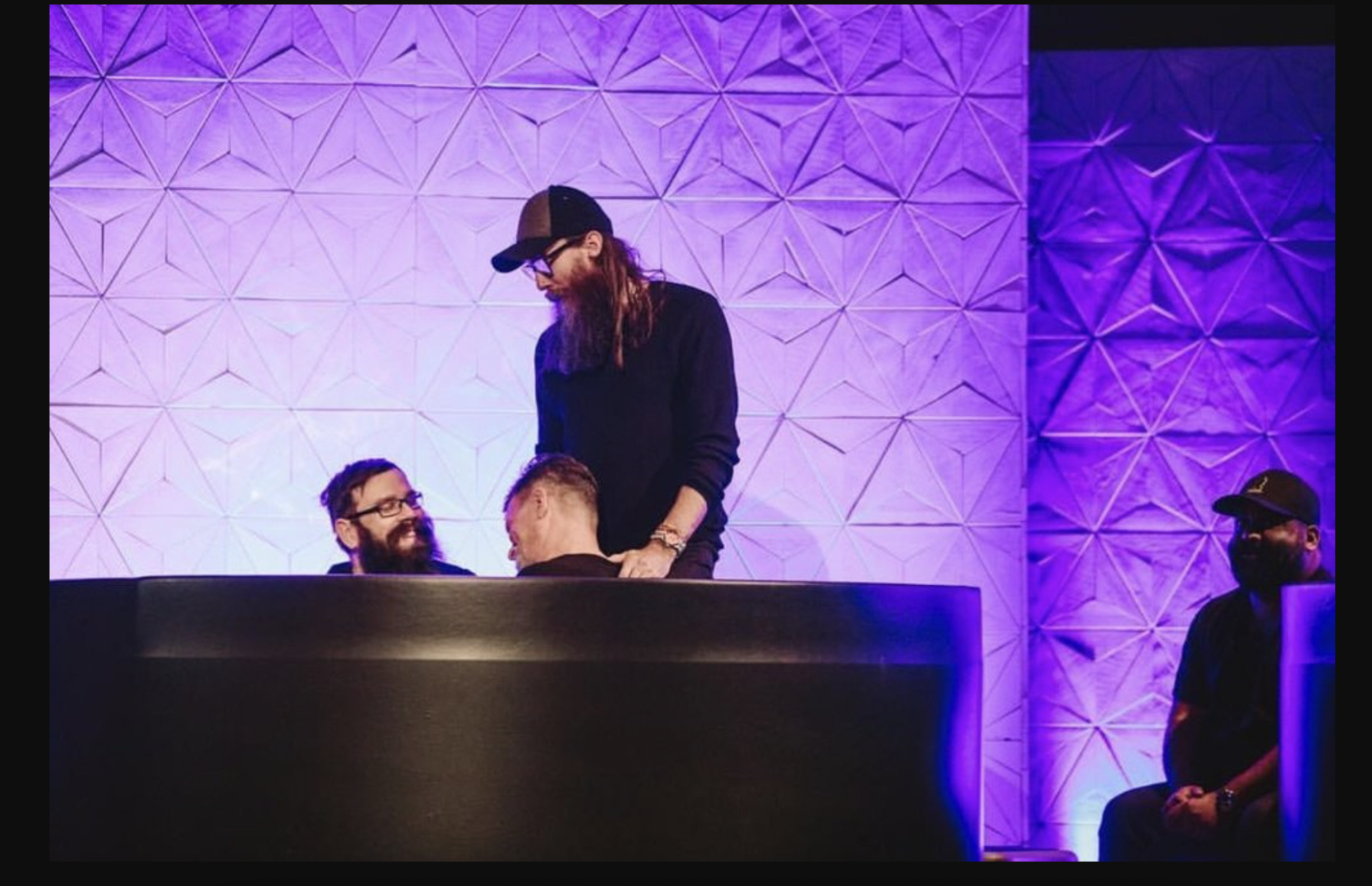 Four men in a dimly lit room with purple lighting and geometric wall design, with three men seated and one man standing and leaning over a seated man, all smiling and engaged in conversation.