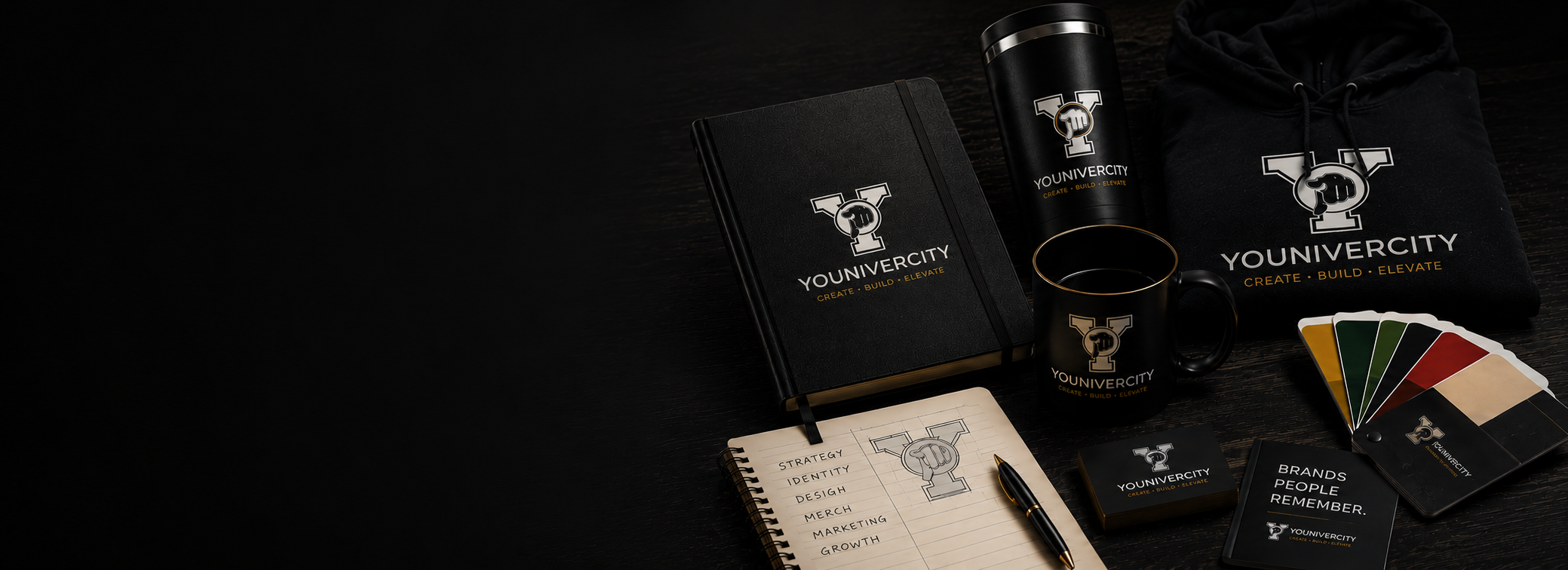 Arrangement of branded office items including a black notebook, a black mug, a black hoodie, color swatches, business cards, a wallet, a black tumbler, a notepad with a handwritten list, and a pen all featuring the Youniversity logo and slogan on a dark surface.