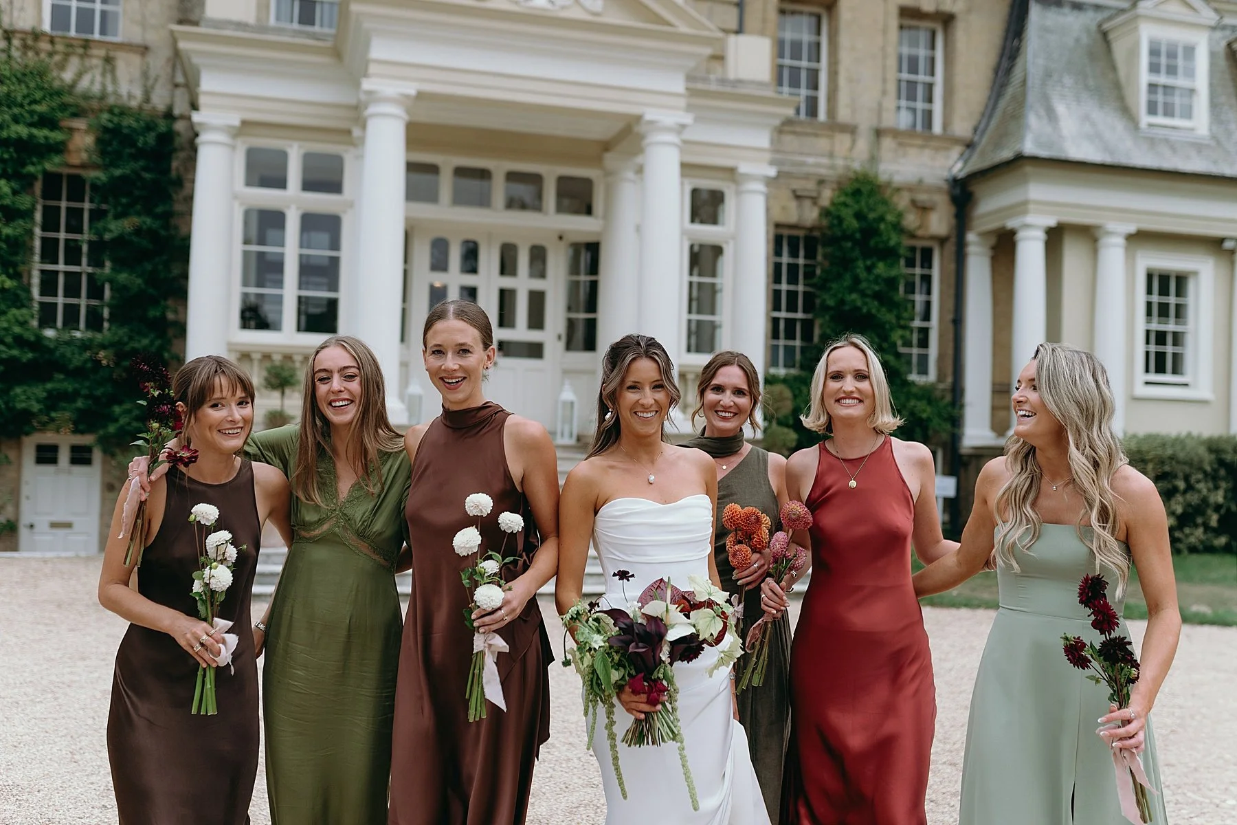 a bride and bridesmaids dressed in autumnal colours are walking and looking towards the camera