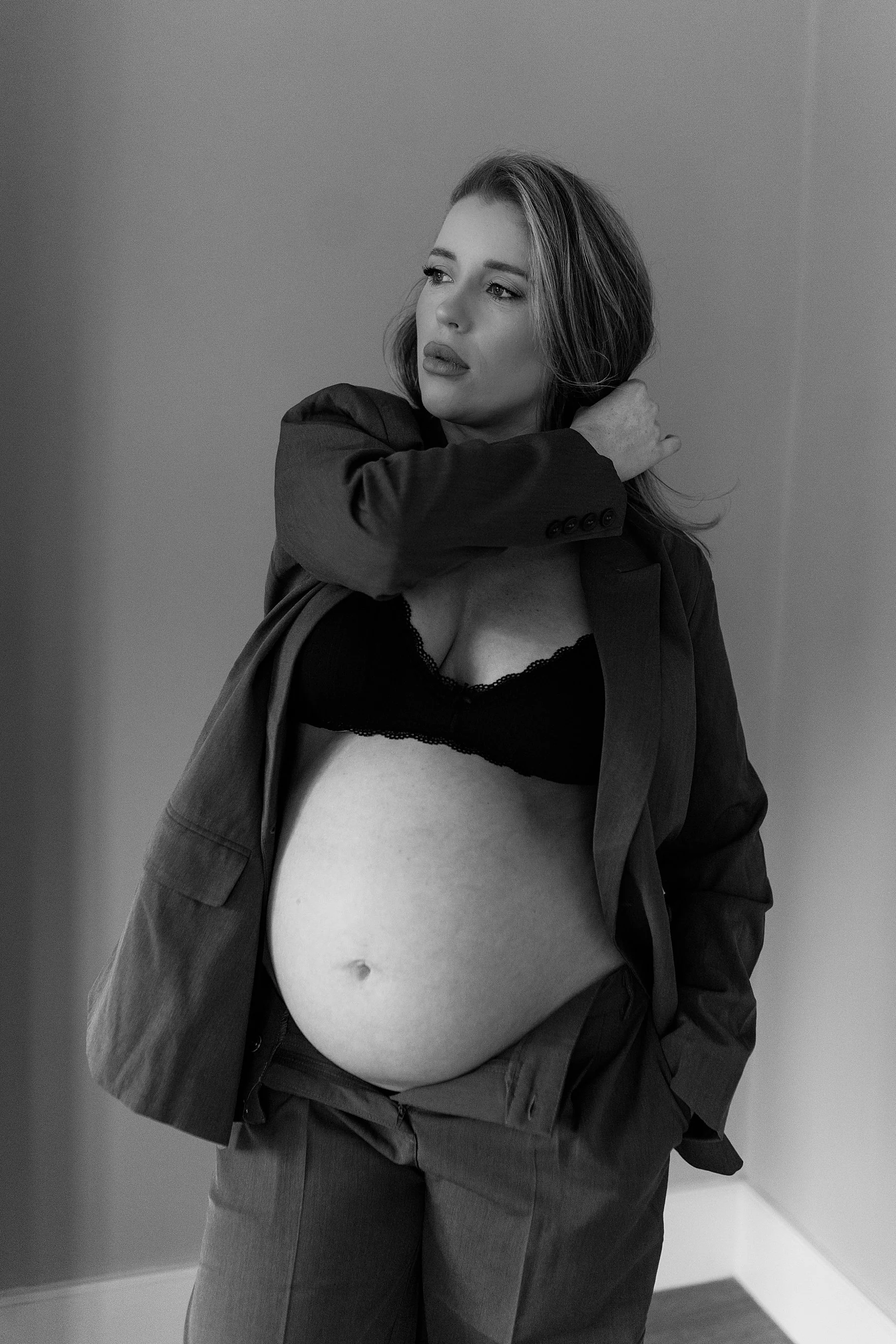 A pregnant woman wearing a black lace bra and a blazer with jeans, standing indoors against a plain wall, looking thoughtful with her left hand touching her neck.