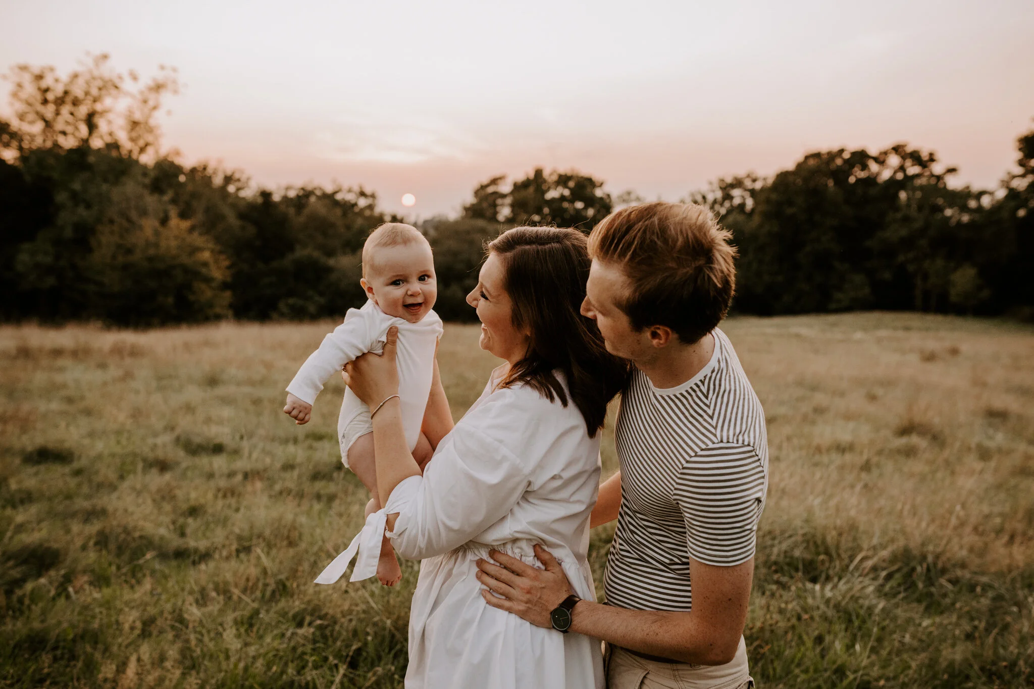Family of three, including a smiling baby held by a woman, standing in a grassy field at sunset, with trees in the background.