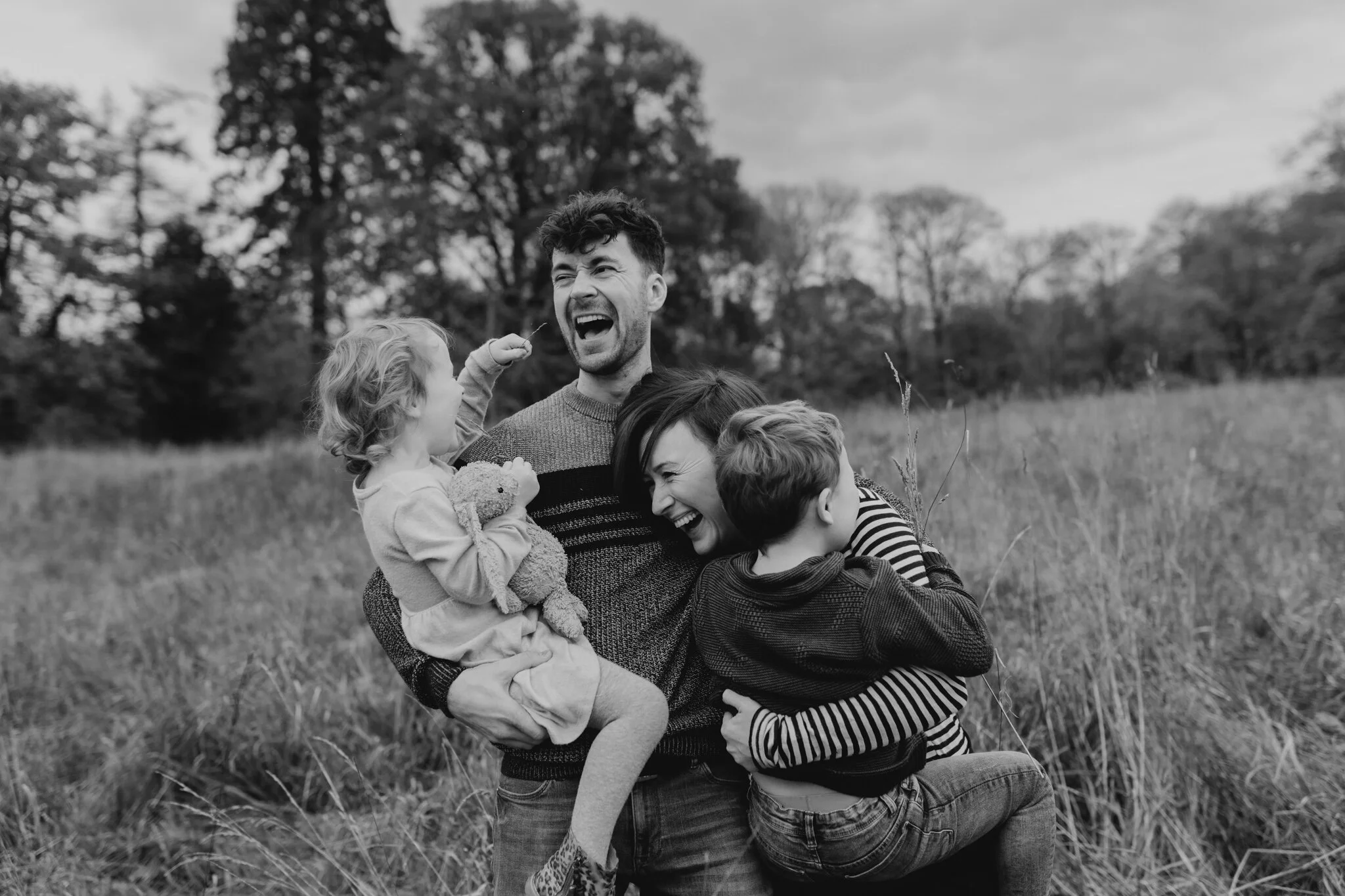 A happy family outdoor in a field, with two children and two adults, all laughing and hugging.