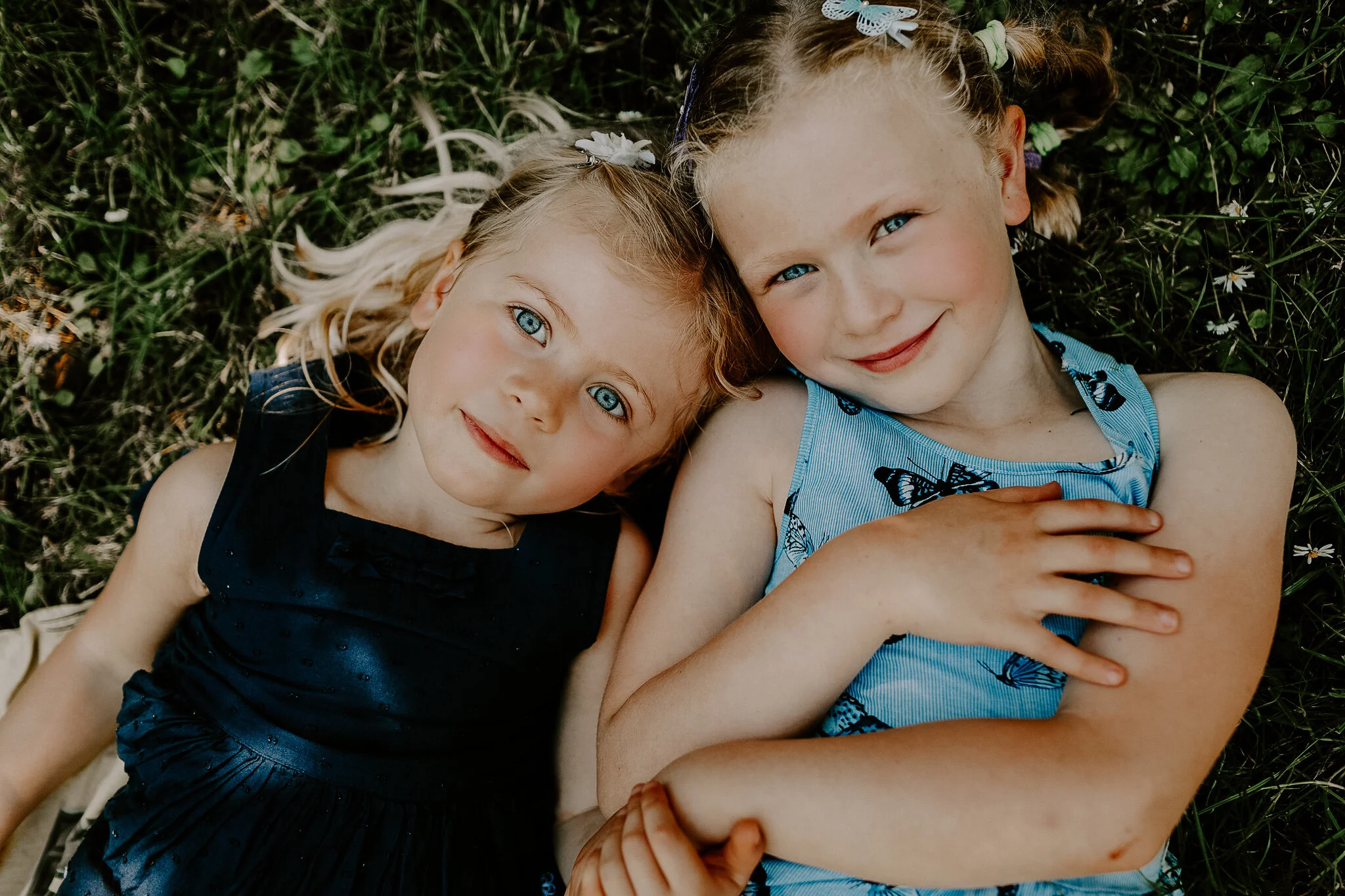 Two young girls with blue eyes lying on grass, one wearing a dark blue dress with butterflies, the other in a blue dress with black butterflies, both with braided hair and ribbons, smiling at the camera.