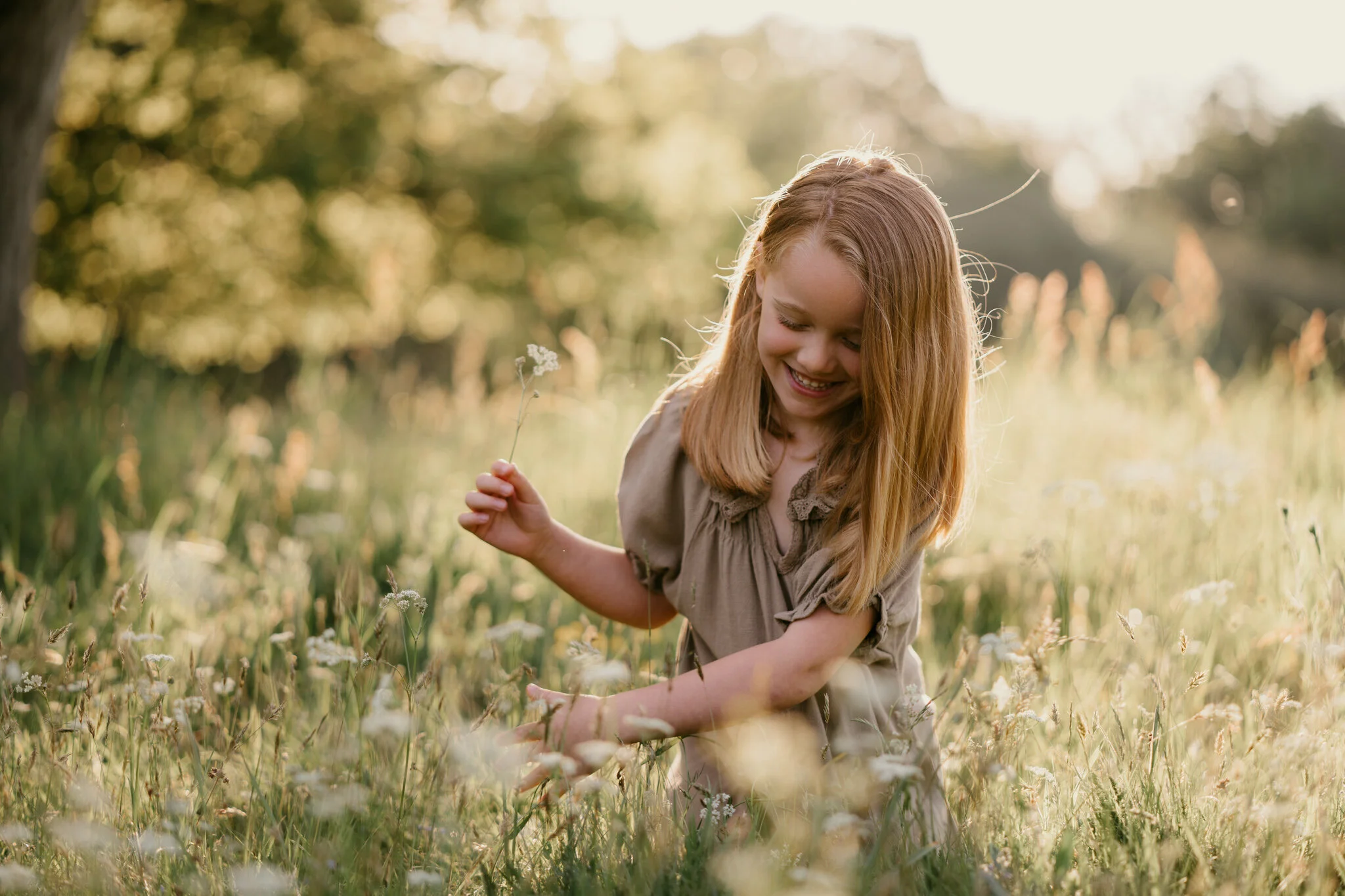 A young girl with red hair smiling and playing in a grassy field with wildflowers during sunset.