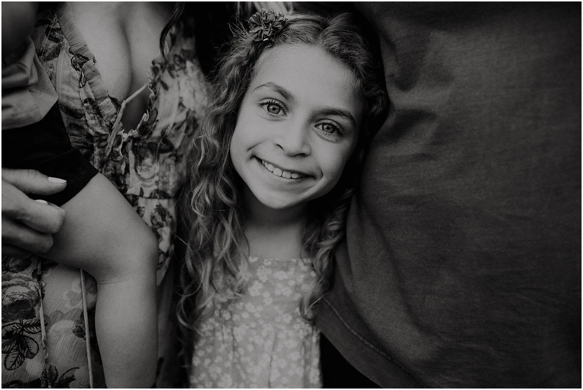 A smiling young girl standing between two adults with her head resting on one person's arm, looking up at the camera.