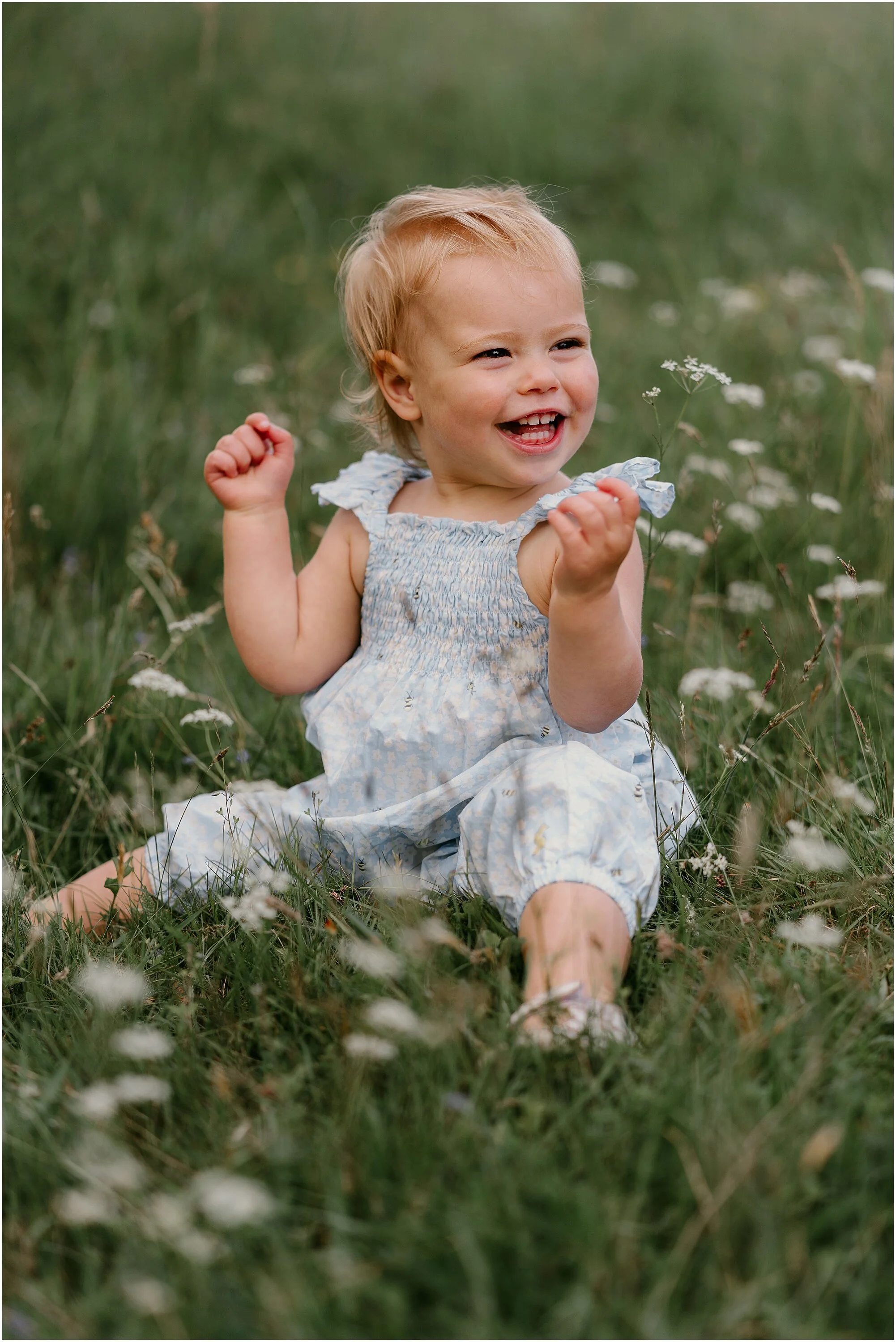 A young girl with blonde hair sitting on grass in a field of small white flowers, smiling and holding a flower in her hand.