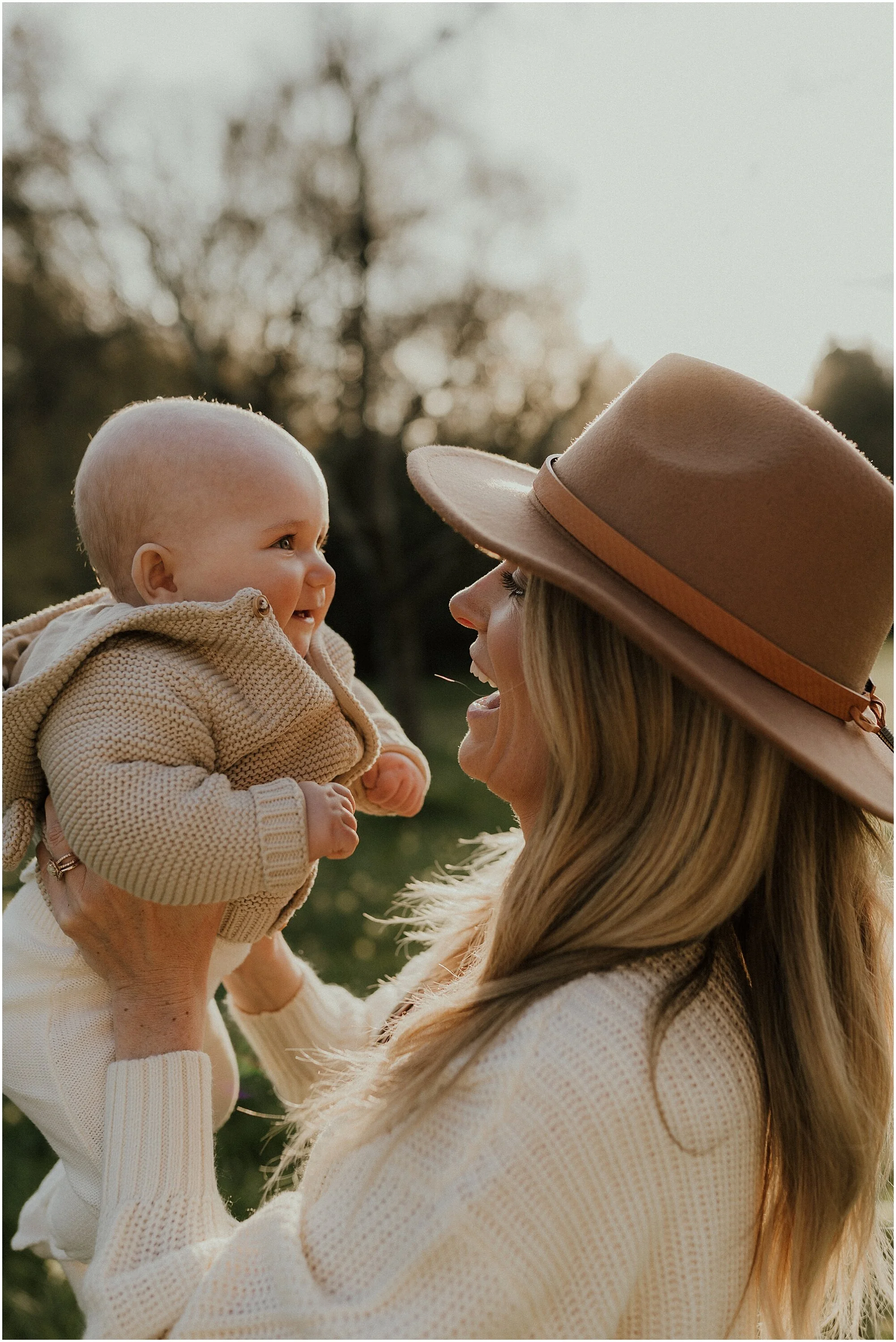 A woman with long blonde hair wearing a large tan hat and a cream sweater holding a smiling baby with a bald head and beige knit sweater outdoors during sunset.