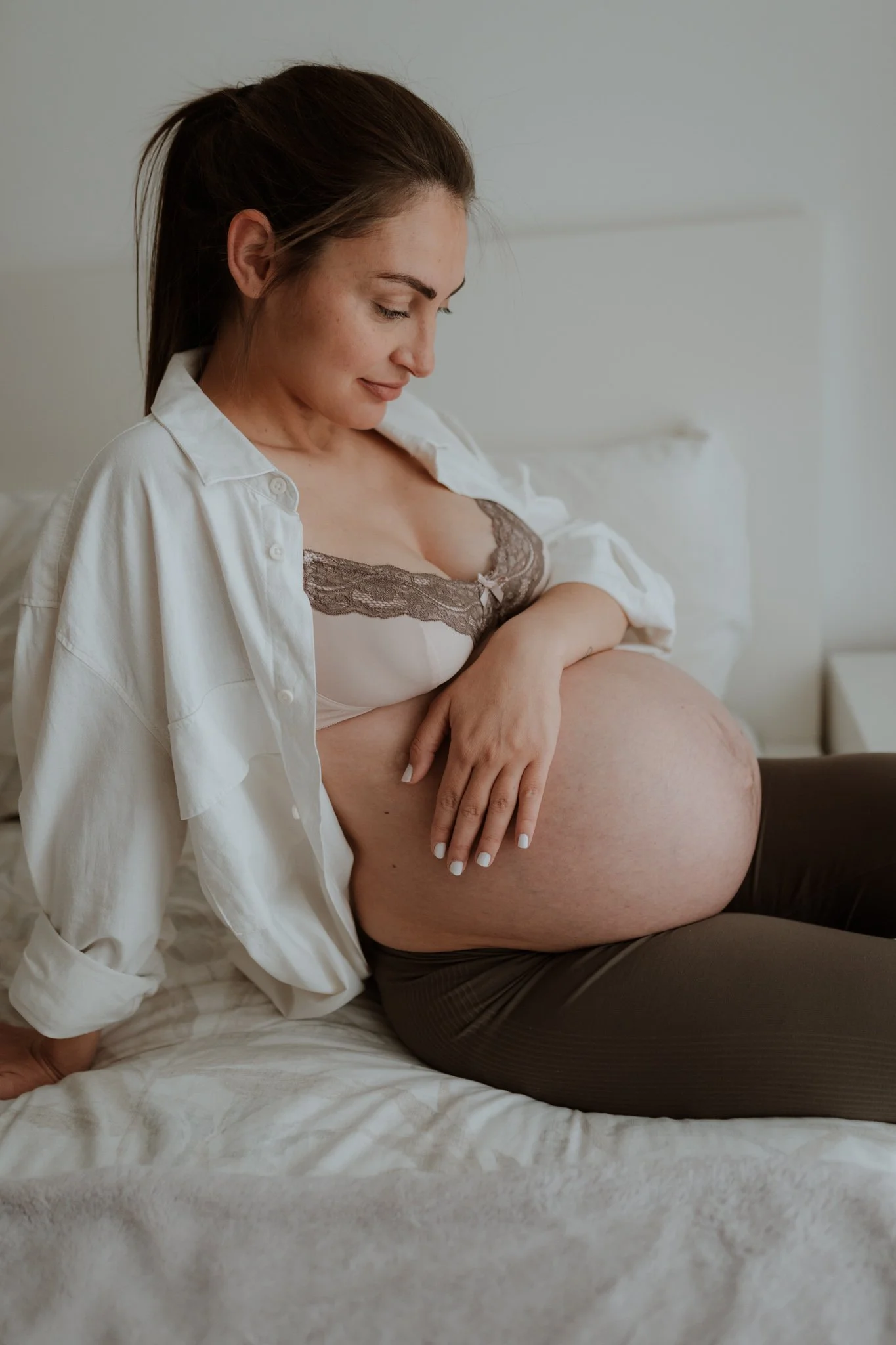 Pregnant woman sitting on a bed, gently touching her large belly, smiling softly, in a cozy bedroom setting.