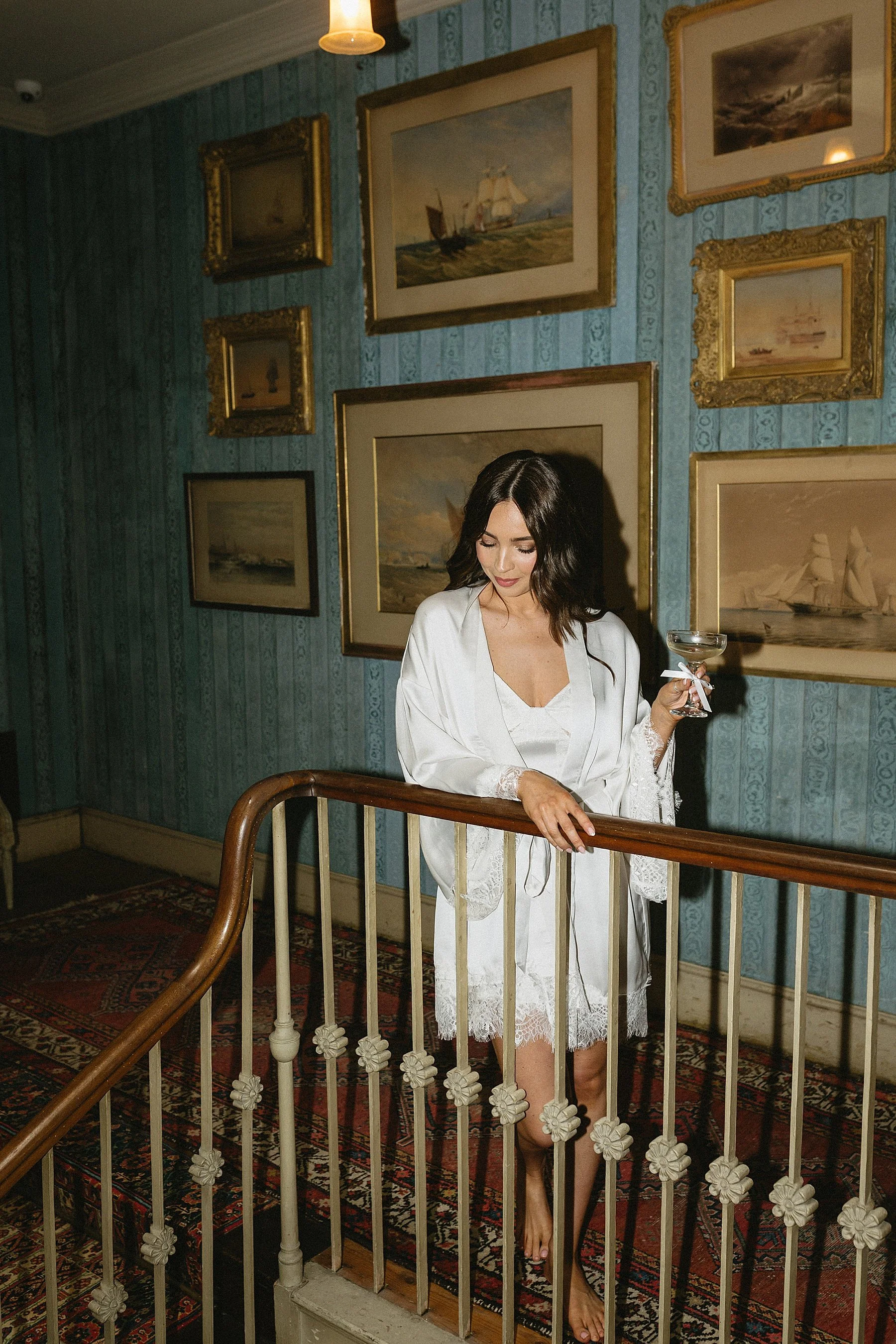 a bride wearing a whote robe stands near. the staircase of her wedding venue, st giles