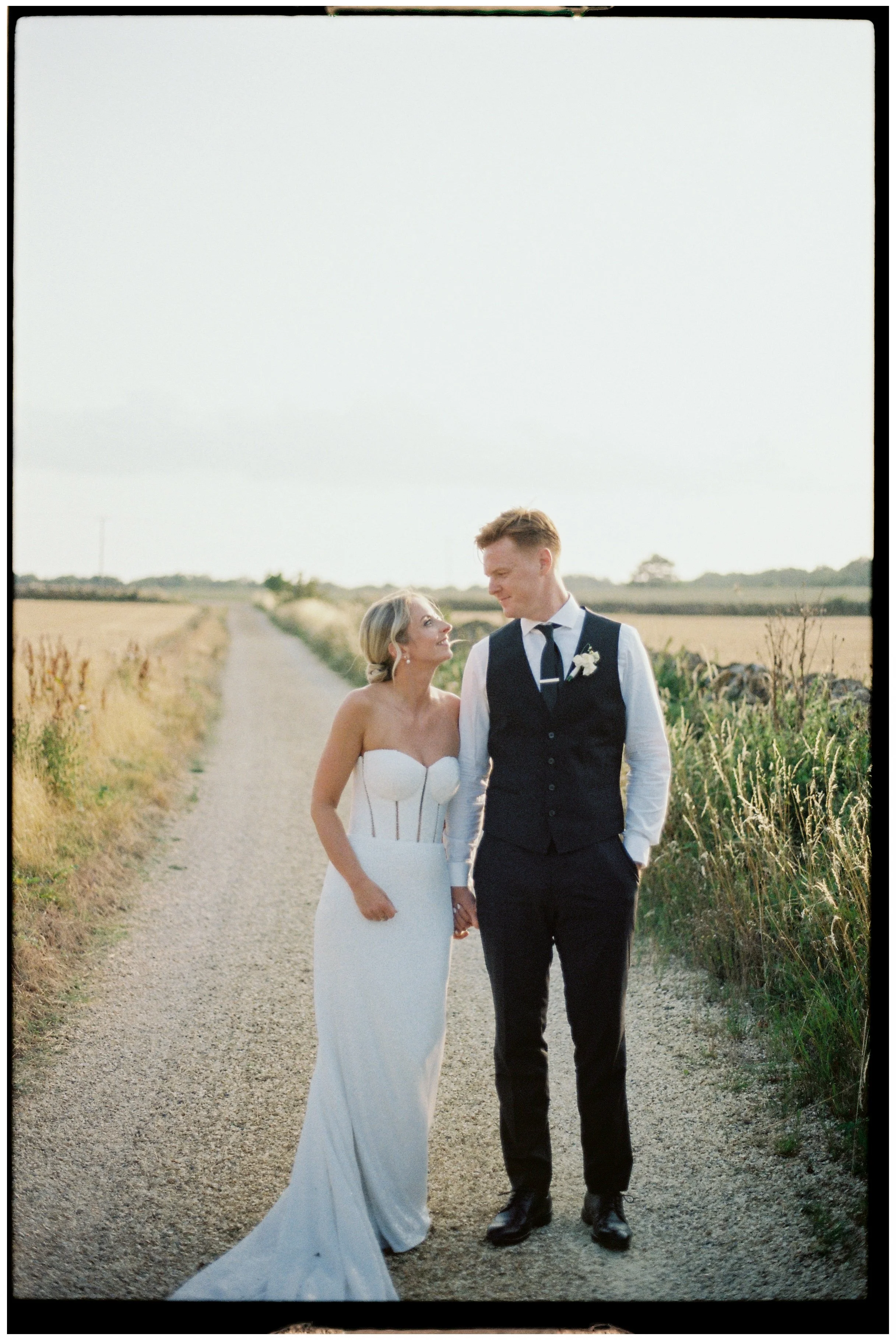 Bride and groom holding hands on a gravel path in a rural field, with the bride in a white strapless wedding dress and the groom in a dark vest and pants, gazing at each other.
