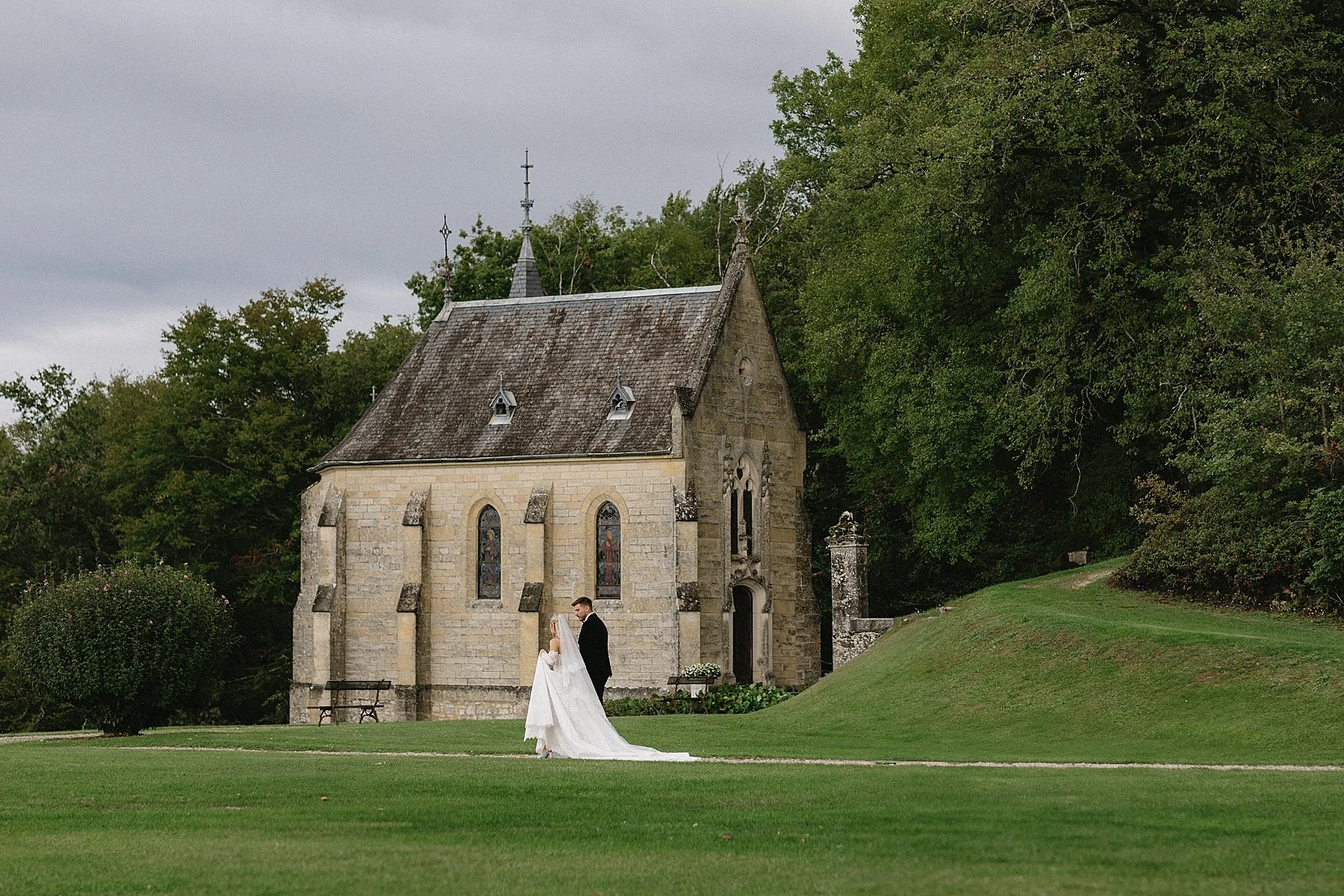 a bride and groom walking towards a french chapel. The background is green countryside