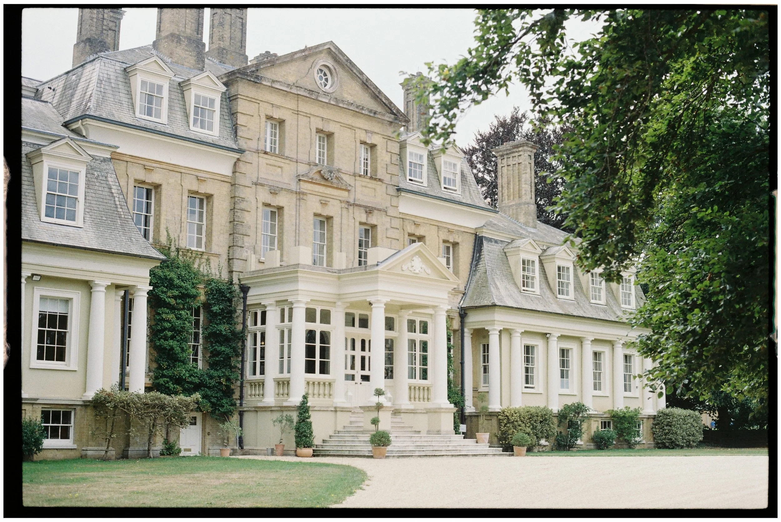 Pylewell Park, a large historic mansion with light-colored stone and white accents, surrounded by greenery, with potted plants and a gravel pathway in front.
