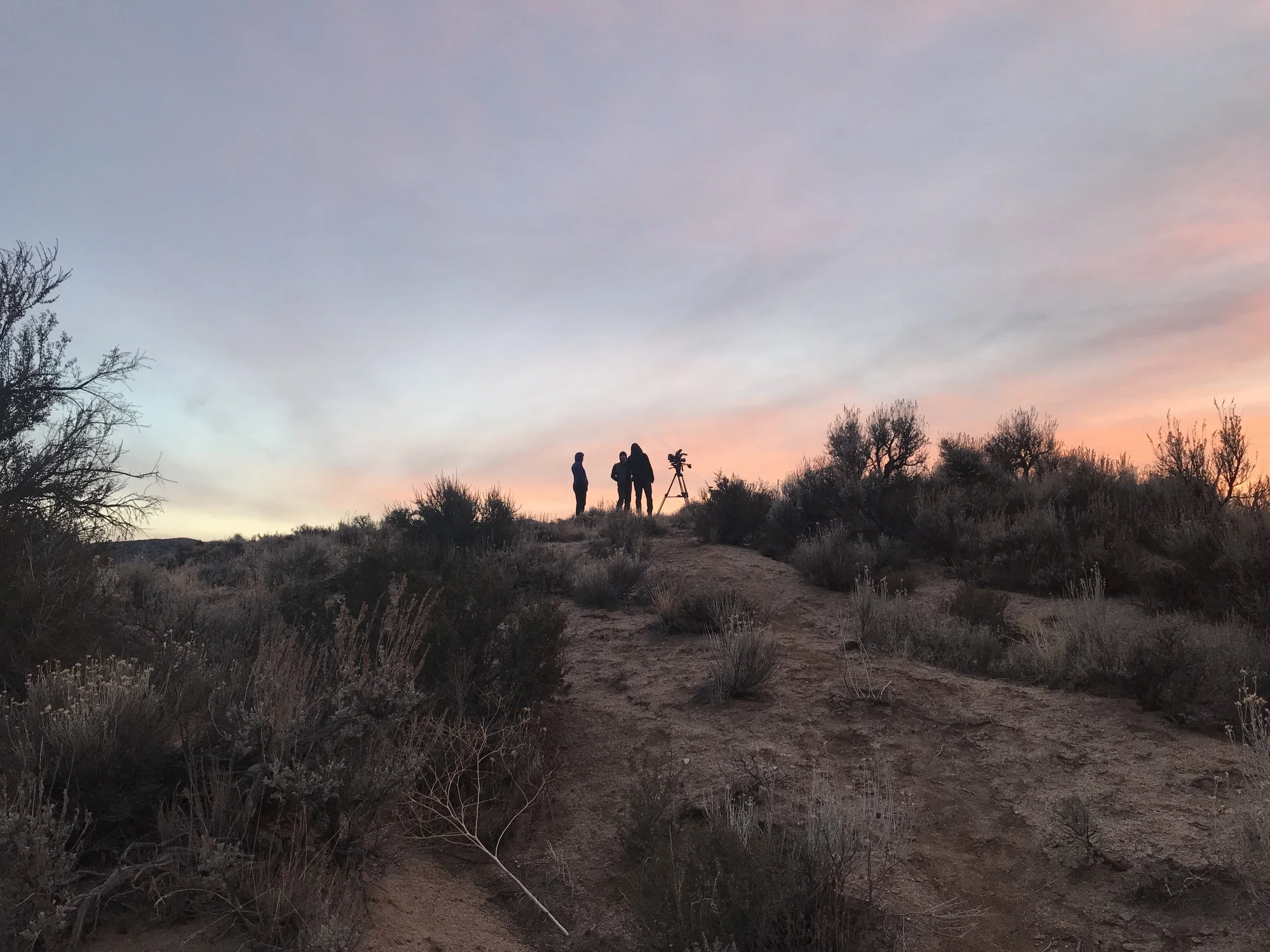 Three researchers with a telescope on a hill at sunset, surrounded by desert shrubs and trees.